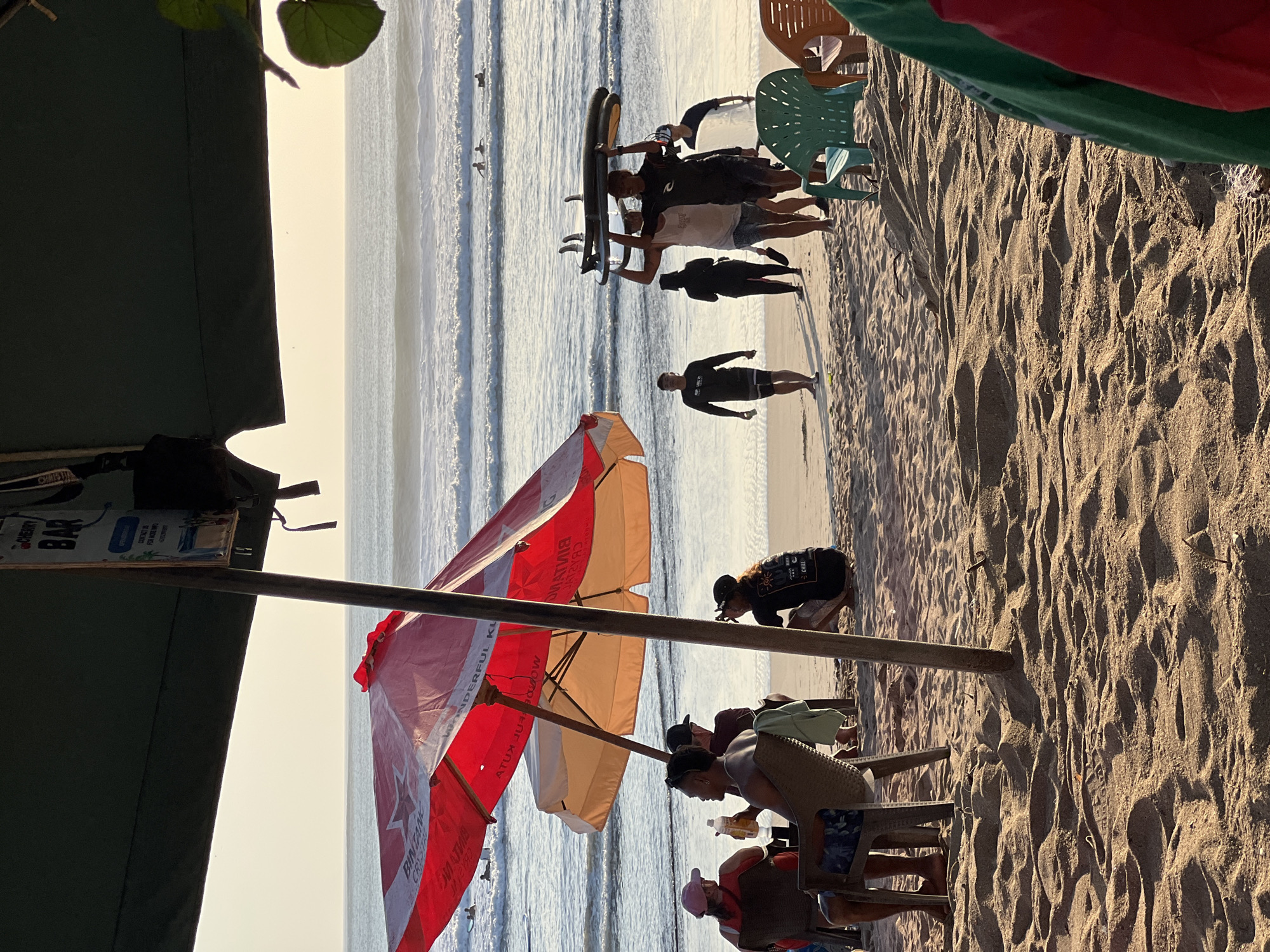 A group of people are gathered on a sandy beach in Kuta, Indonesia, near the ocean. In the foreground, several people are seated at tables under a large red and orange umbrella, some with drinks. Further back, a group of people are walking on the beach, two of whom are carrying surfboards above their heads. In the background, the ocean has gentle waves, and several surfers are visible in the water. The sky is clear and bright, suggesting it is either early morning or late afternoon. A sign for "CHERRY BAR" is visible hanging from an overhead structure.