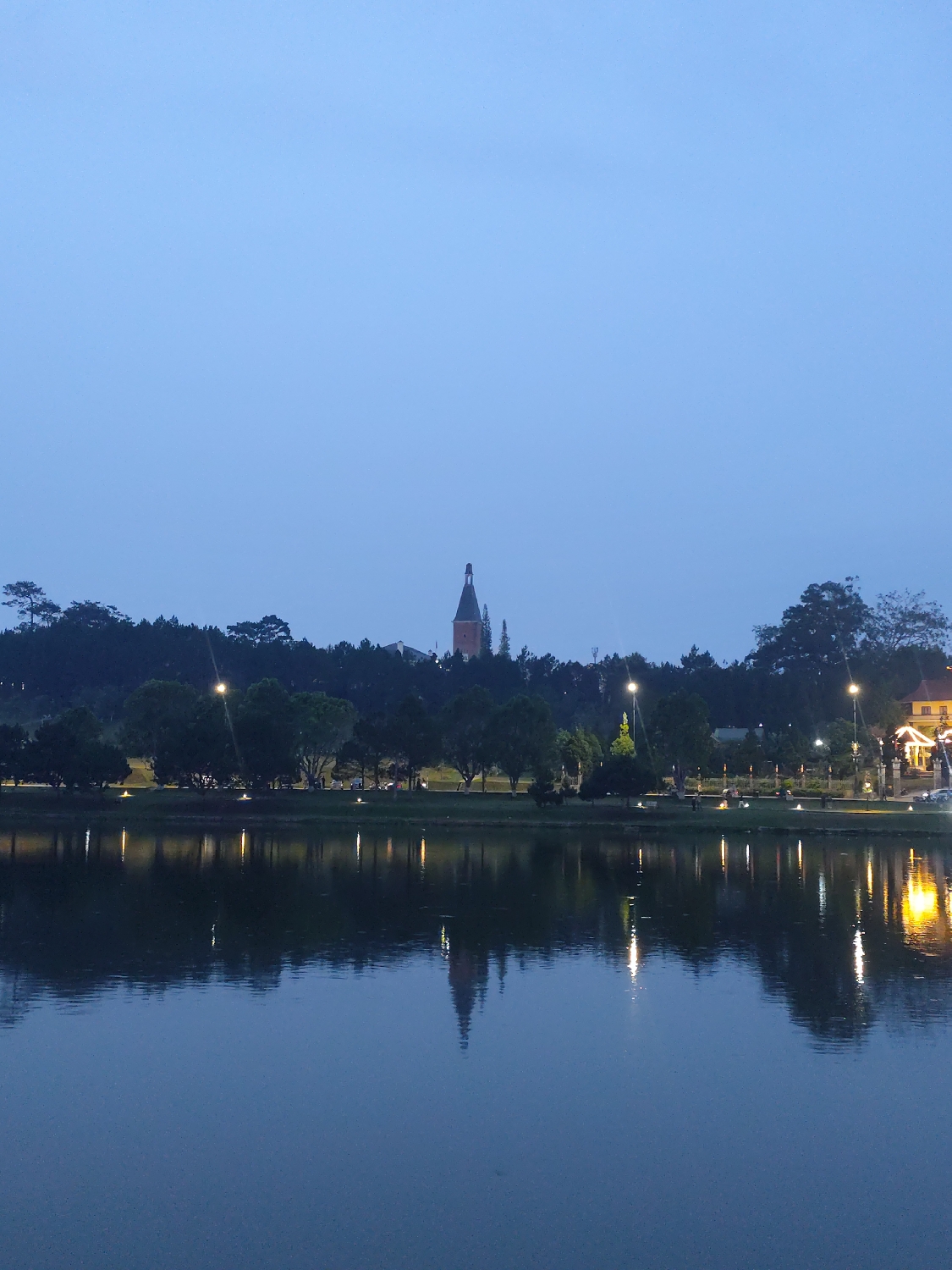 This image presents a serene landscape captured during dusk or early night in Da Lat, Vietnam. The scene is dominated by a calm body of water in the foreground, acting as a mirror for the distant shore. The sky above is a uniform, deep blue, indicating twilight.
Across the water, a dense line of trees forms a dark silhouette along the bank. Behind these trees, a distinctive, spire-topped building stands out against the horizon, slightly to the left of the center. This building's silhouette, along with other faint structures, suggests a distant town. Artificial lights from streetlamps or buildings dot the far bank, casting bright, elongated reflections that stretch across the water's surface. On the far right, a building with warm, glowing lights is prominently visible.
The water's surface is exceptionally still, allowing for clear and almost perfect reflections of both the distant lights and the prominent spire. There are no people or discernible activities taking place, contributing to the tranquil and peaceful atmosphere. The absence of ripples on the water suggests calm weather conditions. No visible text is present in the image. The overall impression is one of quiet beauty, highlighting the reflective quality of the water and the gentle glow of lights against the darkening sky, characteristic of an evening in Da Lat.