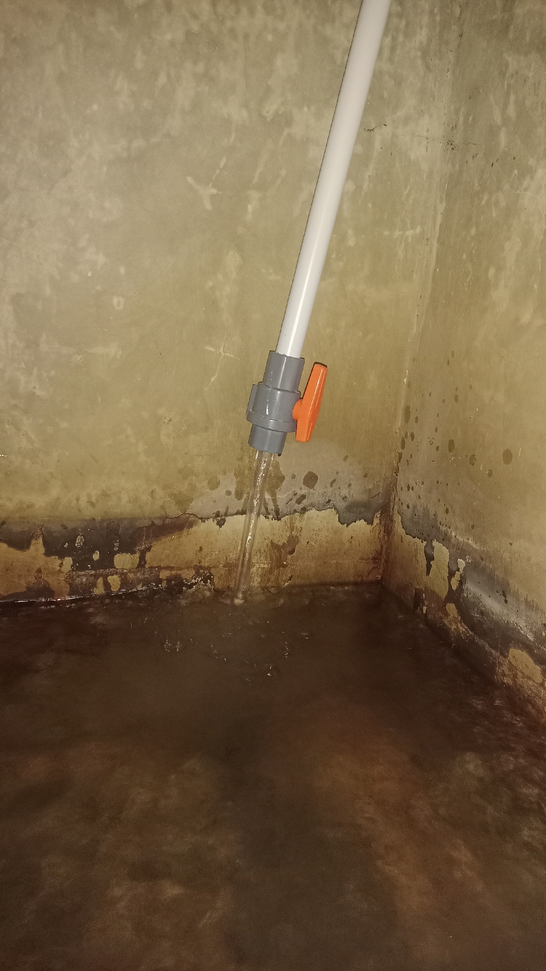 In Desa Burujul Jaya, Indonesia, a corner of a room is depicted with stained, textured walls and a dark, murky floor. A white pipe with a grey and orange valve is angled downwards, with a stream of clear liquid pouring from it into the floor. The valve is open, indicating water is being released. The walls appear to be concrete or a similar material, showing signs of age and moisture damage, with dark, uneven staining near the floor. The overall impression is of a functional, albeit somewhat dilapidated, area where water is being drained or supplied.