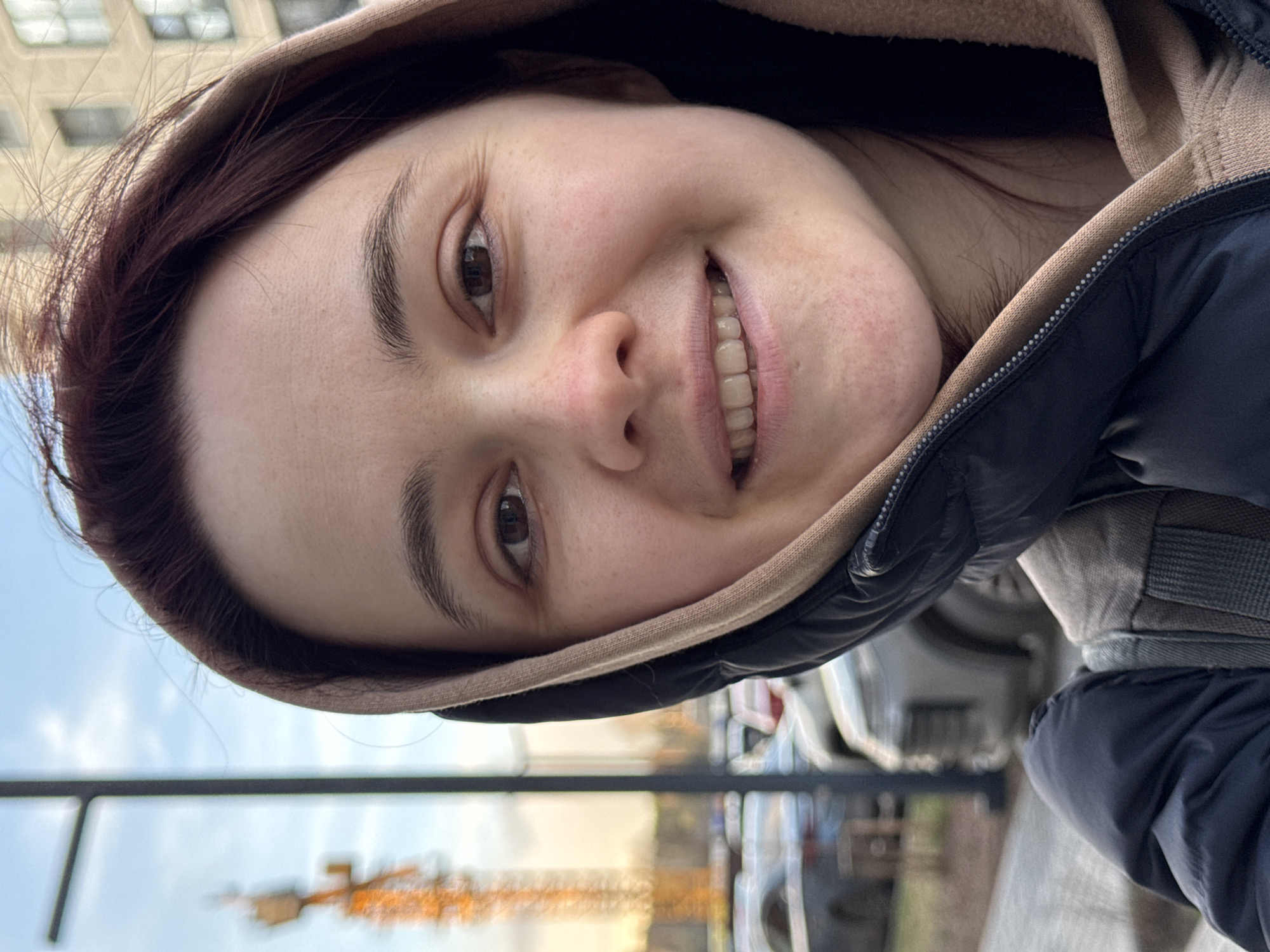 A young adult woman is featured in a close-up, sideways-oriented selfie, looking directly at the camera with a broad, genuine smile that reveals her teeth. She has dark, possibly reddish-brown hair, and dark eyes. Her skin has natural texture, with visible pores and slight freckling. She is wearing a dark-colored, likely black or navy blue, puffer jacket or coat, with a light brown or beige hooded sweatshirt underneath, or as a lining to her main jacket's hood. A zipper line is visible along her clothing.
The image was taken outdoors during the daytime. The background is blurred, indicating an urban setting. Visible elements include the light blue sky, suggesting clear weather. Several buildings and vehicles are vaguely discernible, along with what appears to be a large, yellow or orange construction crane in the mid-ground. Other blurry details hint at street furniture or railings and some distant foliage.
The woman's joyful expression is the central emotional cue of the photograph. The location context provided indicates this scene is in Izhevsk, Russia. There is no visible text in the image.