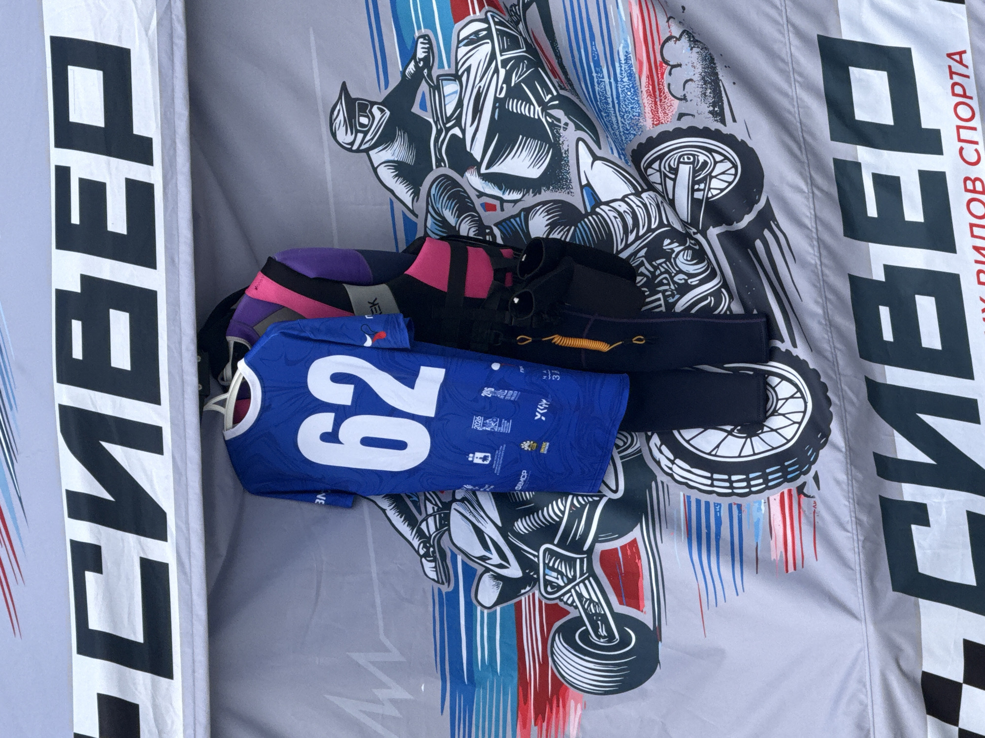 The image shows a section of a gray tent with a printed design of stylized motorcycles and riders. The design features dynamic lines in red, white, and blue, suggesting speed and movement. Two t-shirts and what appears to be a wetsuit or racing suit are hanging on display in front of the tent's backdrop. One t-shirt is blue with a large white number "62" on it. The other garment is a multi-colored suit with purple, black, and pink sections. In the background, across the top of the tent, a large, bold, black Russian word is printed. This word also appears at the bottom of the image, below a checkered flag pattern and the Russian phrase "видов спорта" (types of sports). The context suggests this is likely a promotional display for a sporting event or a vendor selling sports apparel, possibly related to motorsports. The image is a close-up, and no specific identifiable landmarks or street views are visible that would pinpoint the exact location within Perm, Russia, beyond what is suggested by the language.