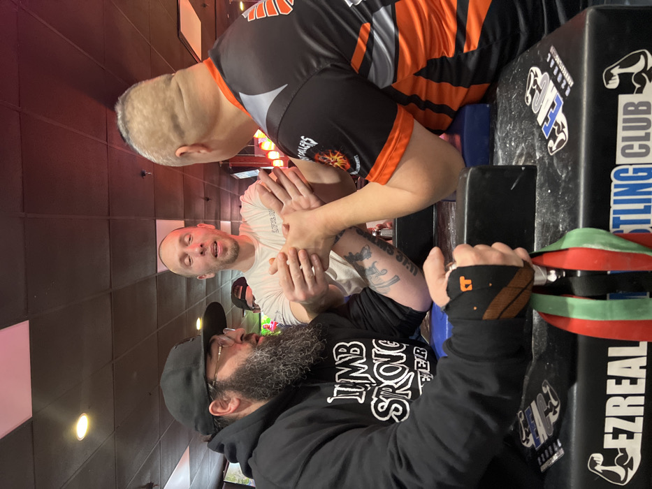 Three men are arm wrestling. Two men are positioned on either side of the arm wrestling table, clasping hands in preparation to begin. One man wears an orange and black jersey while the other wears a white t-shirt. A third man with a beard, wearing a black hoodie and cap, looks on. The arm wrestling table is black with "Wrestling Club" and "EZREAL" written on it. The background includes a black and white checkered wall and some red decorative elements.