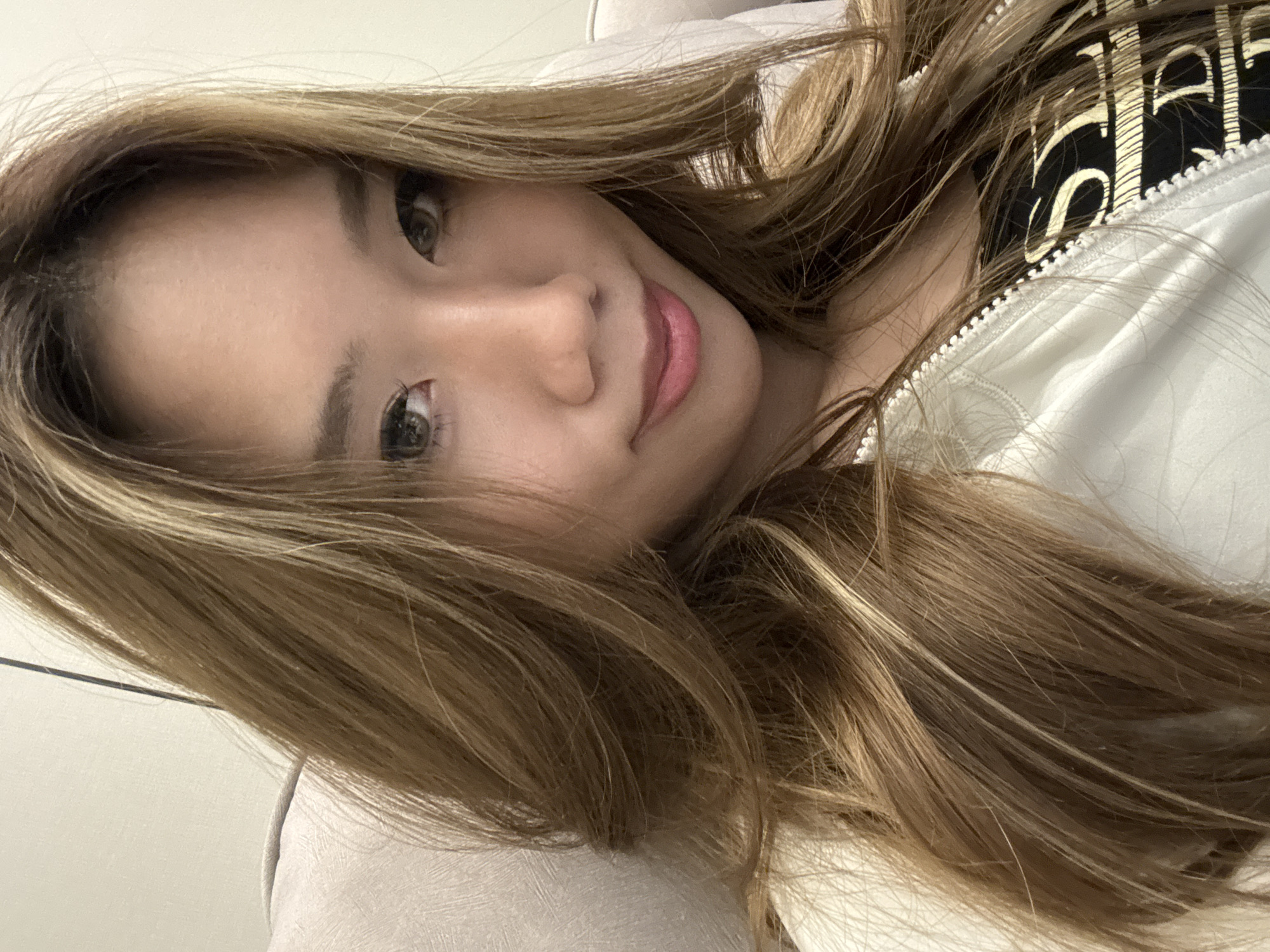 A woman with long, light brown hair is lying on a light-colored surface. She is wearing a white zippered jacket over a black top with gold lettering. Her eyes are brown, and she is smiling slightly. The background is out of focus. The location of the image is not determinable from the given content.