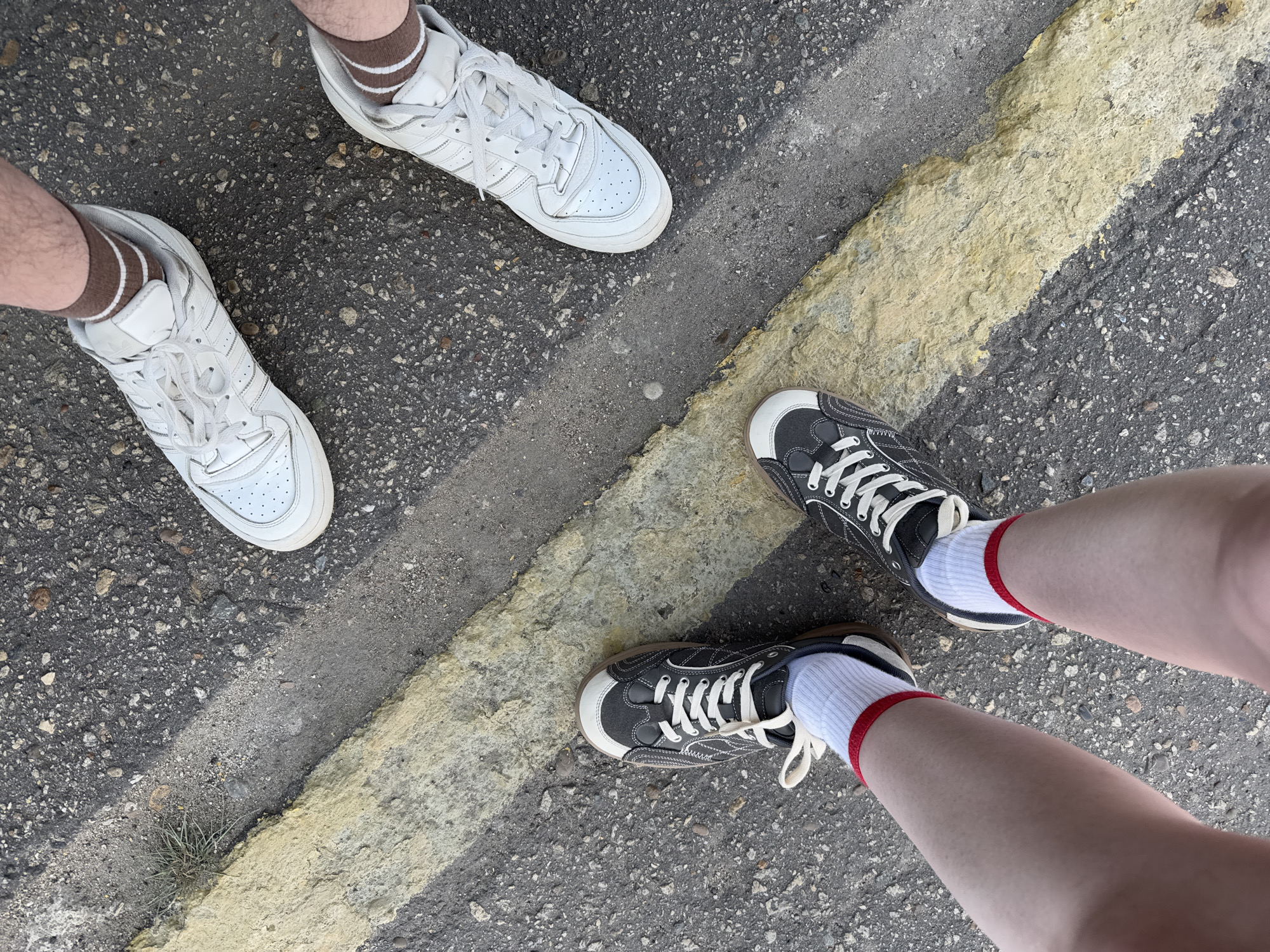 Three pairs of legs and feet wearing sneakers stand on asphalt pavement with a yellow painted line. Two pairs of feet on the left wear white Adidas sneakers and brown socks with white stripes. The pair of feet on the right wear dark gray and white sneakers with white socks that have red stripes. The location and city of Balezino, Russia, cannot be confirmed without further evidence.