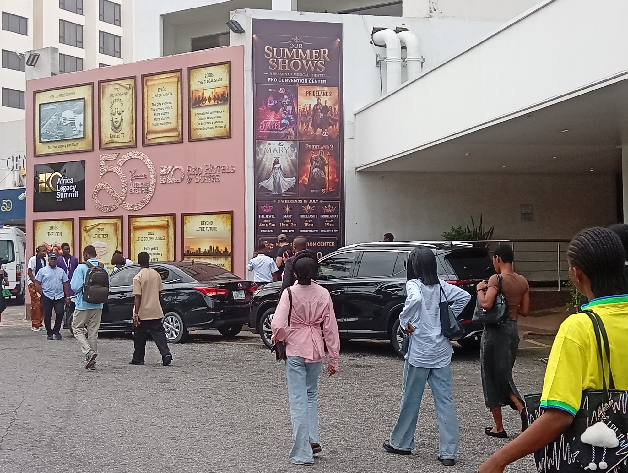 The image shows a group of people walking near the Eko Hotels & Suites complex in Itirin, Nigeria. The group appears to be entering or exiting the Eko Convention Center, indicated by the large posters promoting "Our Summer Shows - A Season of Musical Theatre" and featuring performances such as Jewel, Prideland 2, Mary, and Prideland 3. The posters advertise showtimes for three weekends in July. A large pink sign commemorates "50 years" of Eko Hotels & Suites with historical images. Parked cars are visible nearby and people appear to be walking around them. The location is suggested by the "Africa Legacy Summit" sign as well.