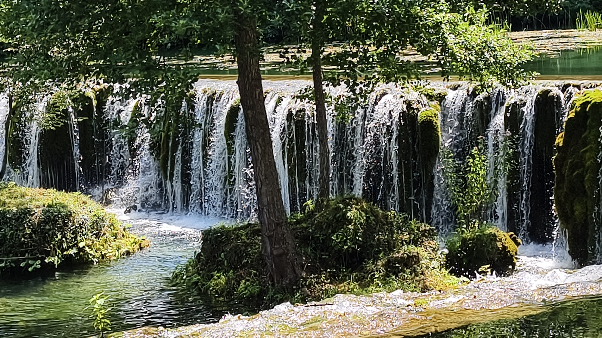 The image shows a cascading waterfall in a natural setting, likely in or near Vrbica, Bosnia and Herzegovina. Lush green vegetation, including trees with dense foliage, surrounds the waterfall. The water flows over moss-covered rocks, creating a frothy, white curtain. The stream in the foreground is clear and reflects the green of the surrounding plant life. There are no people visible in the image, and the scene depicts a tranquil, natural environment. The lighting suggests it is daytime, with sunlight filtering through the trees.