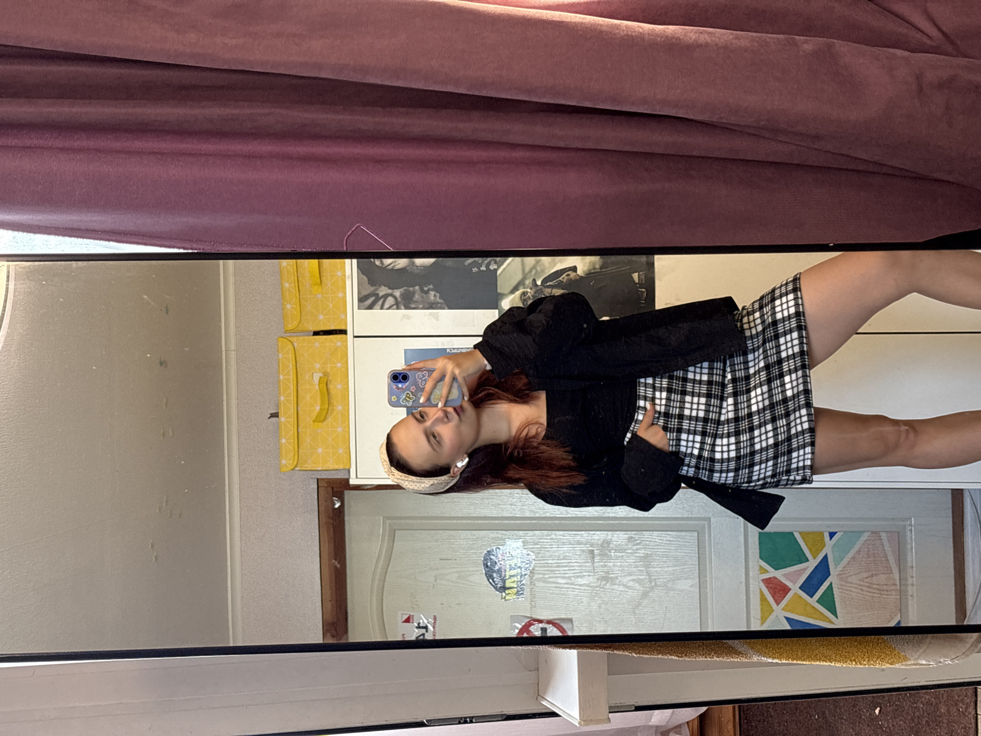 A young woman with auburn hair tied back with a light-colored patterned headband stands in front of a full-length mirror, taking a selfie with her smartphone. She is wearing a black crop top, a black open cardigan, and a black and white plaid mini skirt. She has white earbuds in her ears and is posing with one hand in her skirt pocket, puckering her lips. Reflected in the mirror, behind her, is a white door with various stickers and what appears to be a colorful geometric design. Above the door are two yellow storage boxes with a white pattern. To the right of the mirror are dusty purple curtains. The room's ceiling is visible, showing some imperfections. The overall environment suggests a personal living space, possibly a bedroom or hallway. There is no information to suggest this image is located in the city of Прикамский, Russia; it is likely a domestic setting.