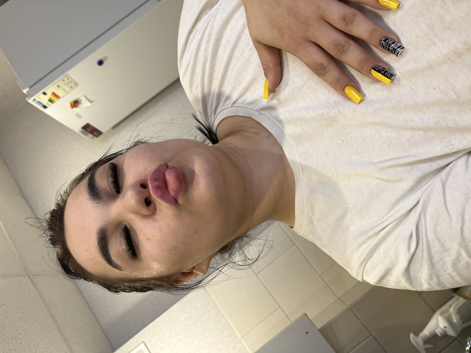 A person with dark hair, pronounced eyebrows, and full lips posed indoors. Their nails are painted yellow with zebra-striped accents on two fingers. They are wearing a white t-shirt. The background includes a tiled wall, a white appliance with stickers, and a glimpse of a white bottle and floor. The location is Chaikovsky, Russia.