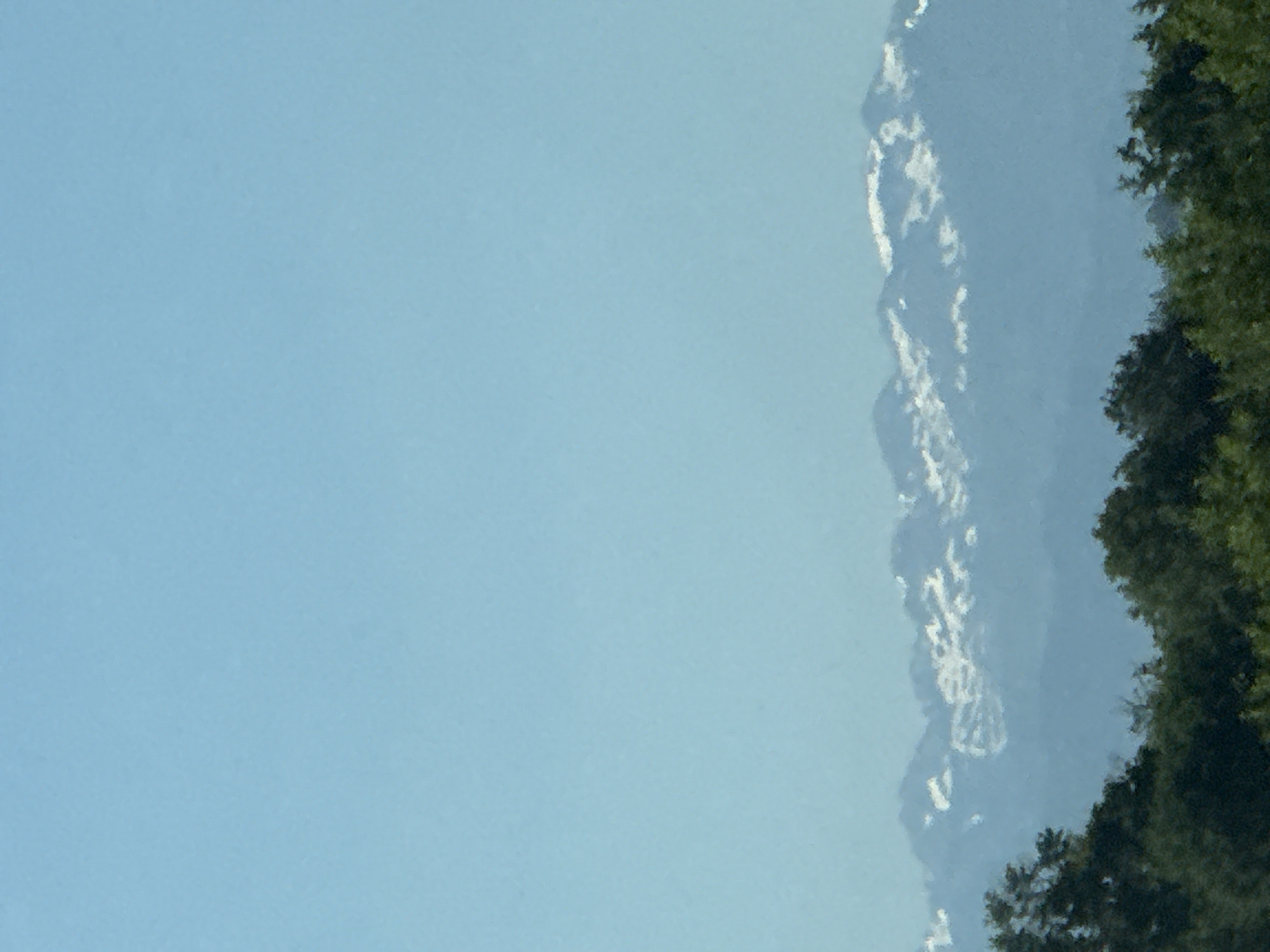 The image shows a landscape with a clear, light blue sky occupying a significant portion of the frame. Adjacent to the sky is a mountain range, partially obscured by haze. The peaks appear to have some snow or ice cover. To the right of the mountains, there are densely forested areas. The image is angled, with the sky appearing on the left. The scene likely captures a mountain vista on the outskirts of or near Saint-Élix-le-Château, France.