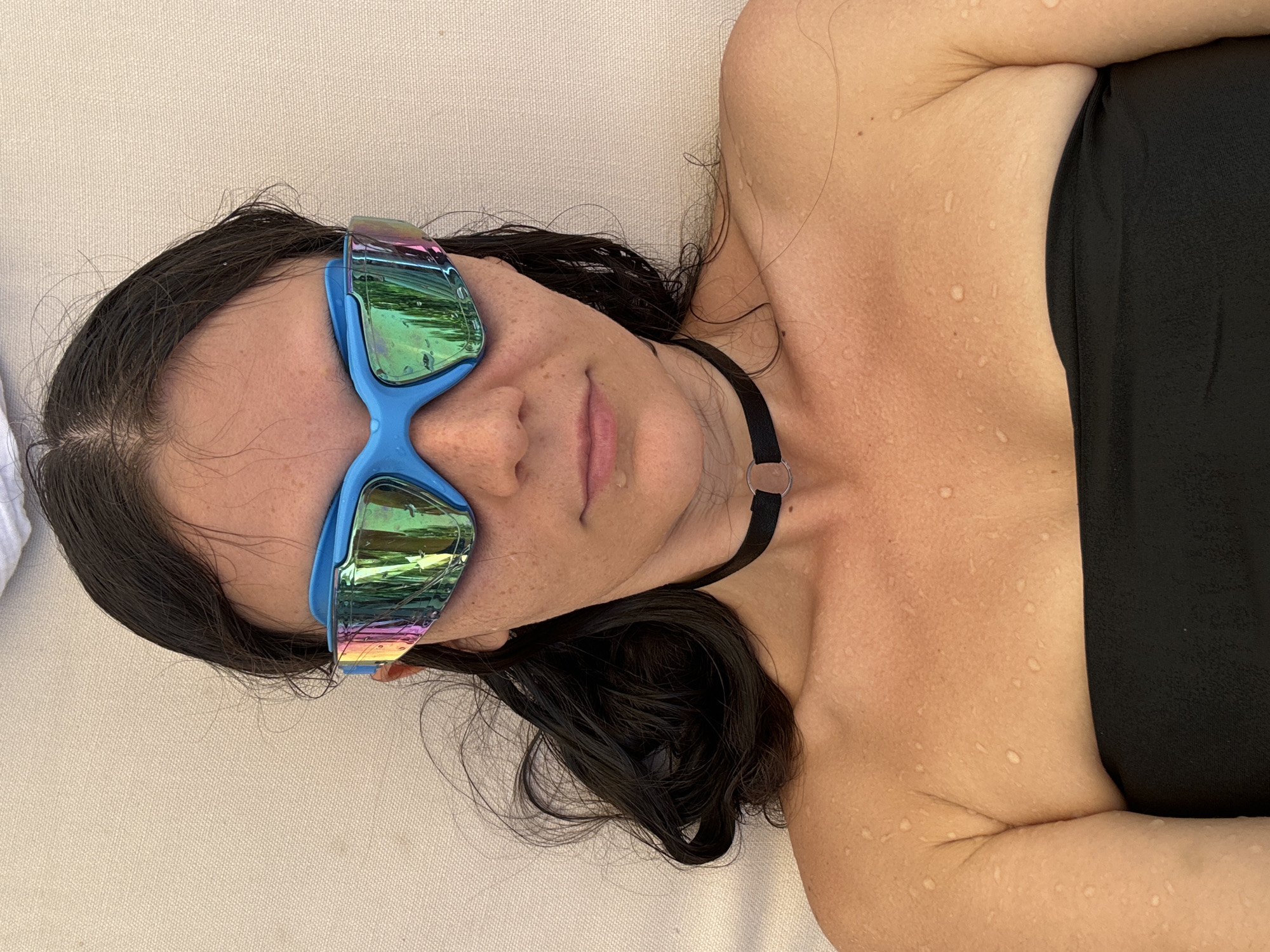 A woman with dark, wet hair lies on a beige surface, likely a lounge chair, in what appears to be an outdoor setting. She wears a black strapless top and a black choker necklace with a circular pendant. On her face are blue-framed swimming goggles with reflective lenses. The woman's skin is fair and has water droplets on it, suggesting she has recently been swimming or exposed to water. The scene is likely located in the city of Phu Thuy Ward, Vietnam.