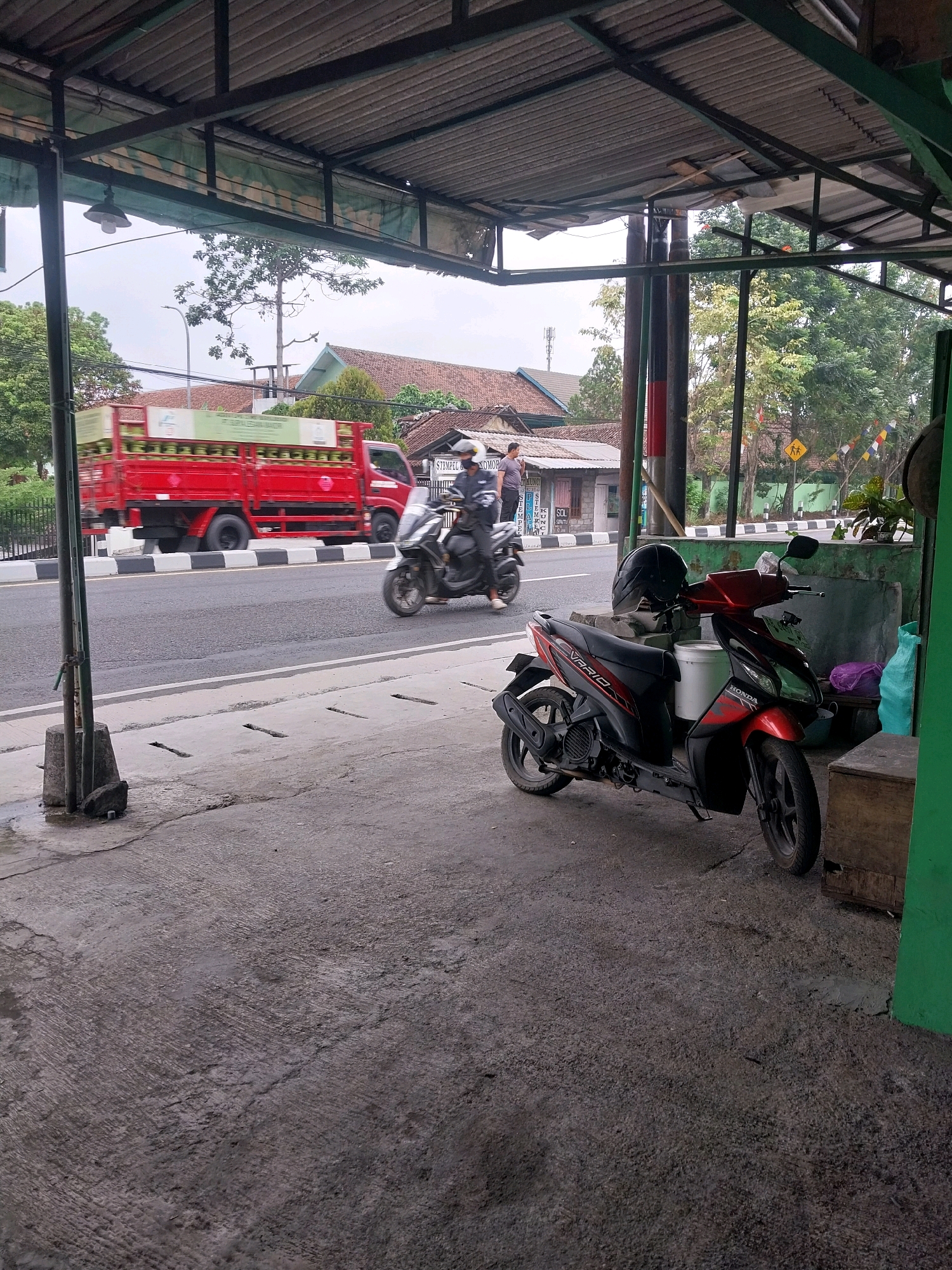 This image captures a scene on a roadside in Sleman, Indonesia. In the foreground, a red scooter is parked on a concrete surface under a corrugated metal roof supported by metal poles. A black helmet rests on the scooter's handlebars. To the left of the scooter, a dark, rough-surfaced concrete area shows some puddles, suggesting recent rain or a damp environment.
In the middle ground, a red truck loaded with what appear to be gas canisters is driving on the road. The truck has a white stripe and writing on its side, but the text is not clearly legible. A motorcycle with a rider is also moving on the road. Two people are standing near a building on the left side of the road. The building has a red tile roof and white walls.
In the background, trees and more buildings with red tile roofs are visible. A yellow pedestrian crossing sign is present on the right side of the road, indicating a crossing point. The sky appears overcast, suggesting a cloudy day. The overall setting appears to be a commercial or residential area along a moderately busy road. The image seems to be taken from a sheltered area, possibly a shop or a food stall, looking out towards the road.