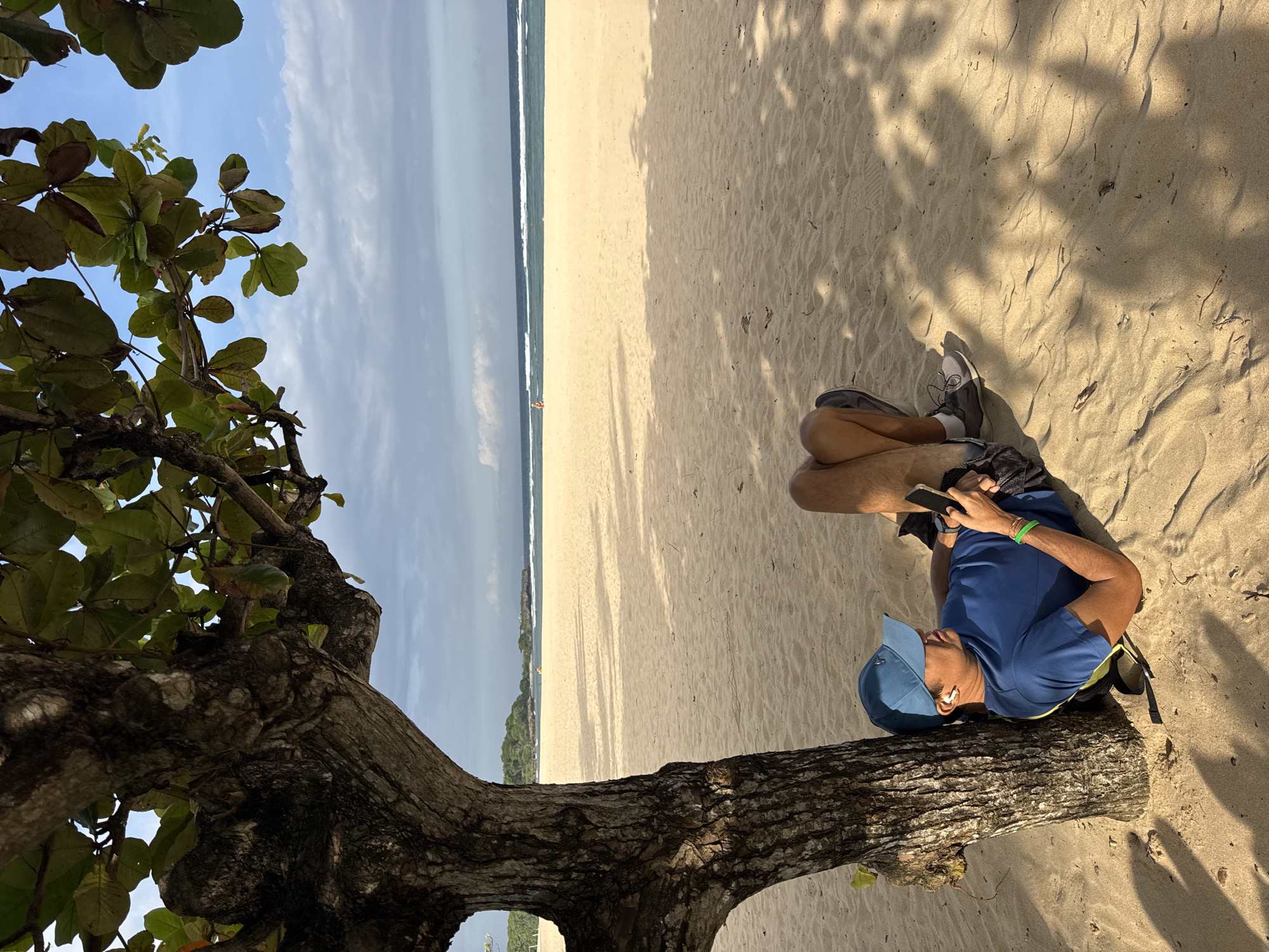 A man in a blue shirt and baseball cap is reclined on a tree trunk on a sandy beach. He is looking at his phone. The beach stretches out to the ocean, with trees and foliage along the shoreline. The sky is partly cloudy. This photograph was taken in Kuta Selatan, Indonesia.
