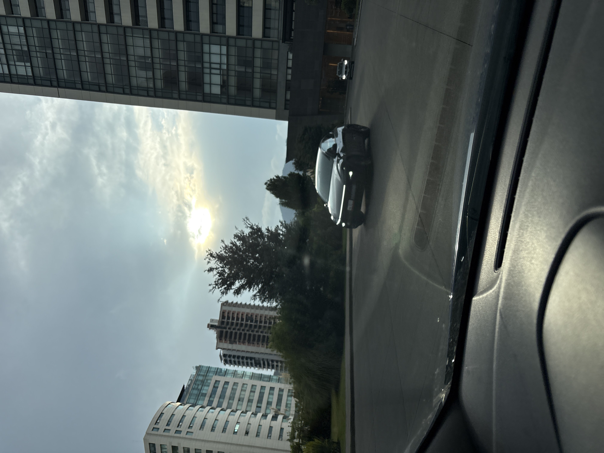 The image shows a view from inside a car, possibly in Jesús del Monte, Mexico, looking out onto a street lined with tall buildings and trees. A dark-colored car is driving on the street in front of the viewer's car. Other buildings are visible in the distance, and the sky is partially cloudy with the sun shining through. The interior of the viewer's car is partially visible in the foreground.