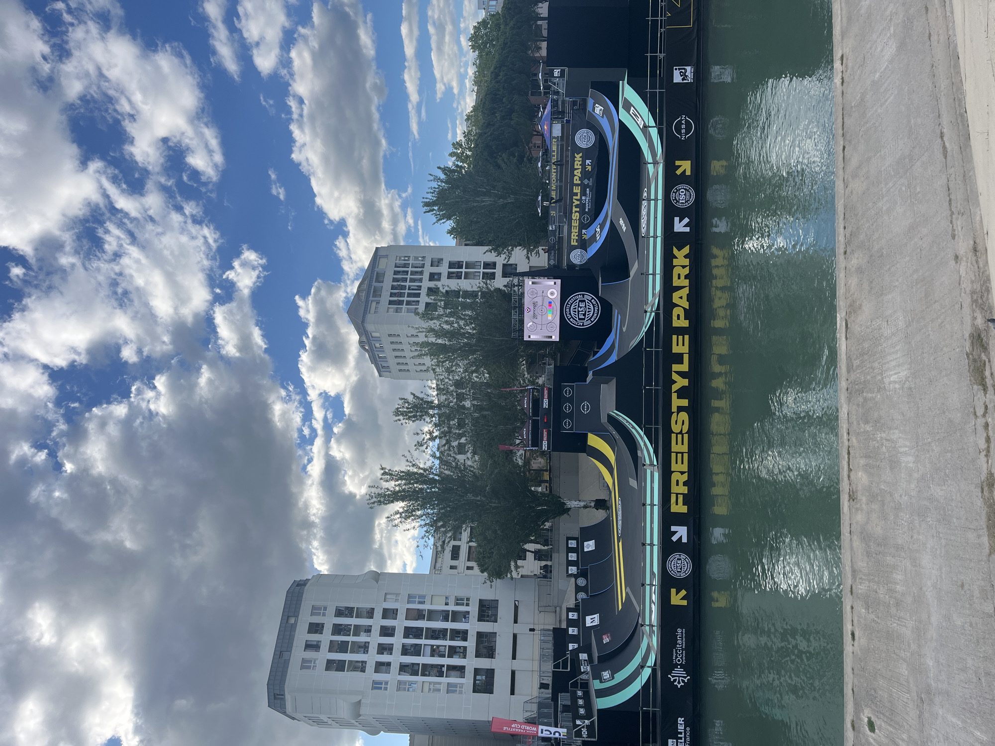 The image displays a freestyle park event, likely for skateboarding or similar sports, in Montpellier, France. The park is positioned adjacent to a body of water, with the event name "FREESTYLE PARK" prominently displayed. The setting includes buildings in the background, and a bright blue sky with fluffy clouds fills the upper portion of the image. Various sponsors' logos, including Nissan and La Région Occitanie, are visible. There appears to be a screen displaying event information or a live feed of the performances.