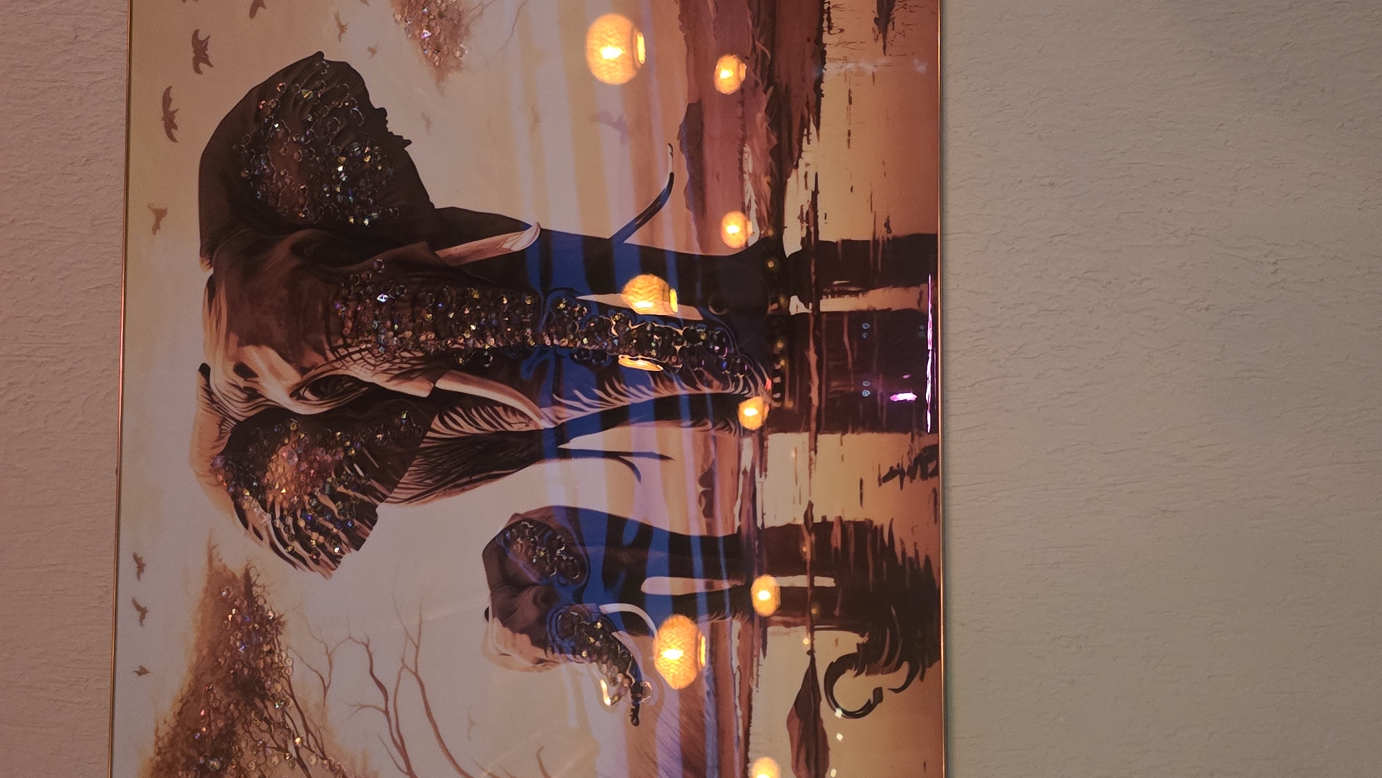 The original image displays a photograph of a piece of artwork, presented rotated 90 degrees clockwise. The artwork itself, when viewed upright, depicts two elephants in a stylized, dreamlike landscape, likely at dusk or twilight.
The dominant feature of the artwork is a large elephant, shown from its head and upper body. Its form is detailed with shades of brown, and its head, ears, and trunk are heavily embellished with shimmering, iridescent sequins or glitter, adding a striking texture and sparkle. Below and slightly to the right of the larger elephant, the head of a smaller elephant is visible, also adorned with similar glittering elements.
The elephants are positioned above a reflective surface, possibly water, which mirrors their forms and the surrounding environment in a blurred, impressionistic manner. The background features a warm color palette of oranges, browns, and purplish-blues, suggesting a sunset or evening scene. Several glowing, orb-like lights, resembling lanterns or fireflies, are scattered throughout the mid-ground and reflected in the foreground, enhancing the ethereal ambiance. Sparse tree branches and small, dark bird silhouettes are visible in parts of the background.
The artwork is enclosed by a thin, gold-colored frame visible on its left side. To the right of the framed artwork, a textured, light-colored wall is visible, indicating the piece is displayed indoors. There is no visible text in the image.