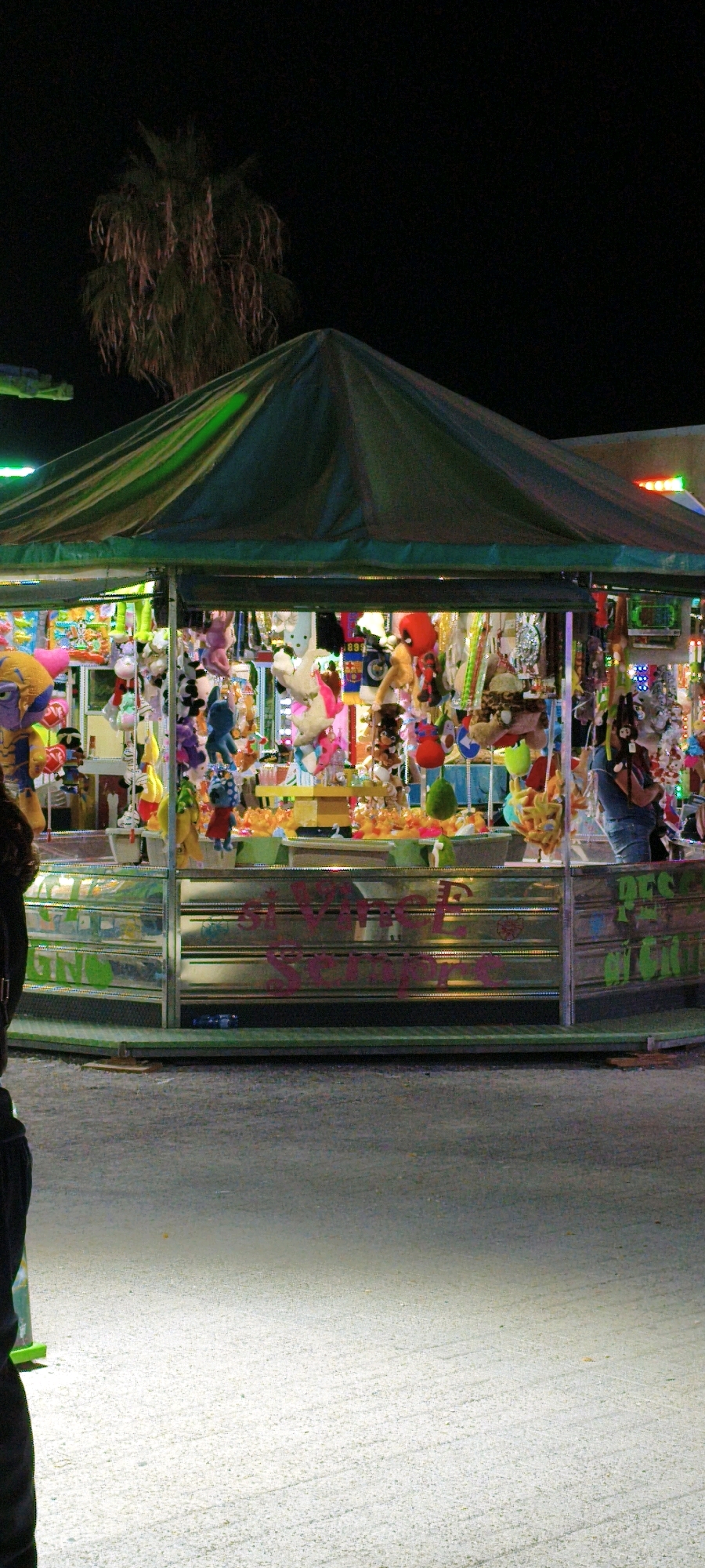 The image shows a brightly lit stall filled with plush toys and other prizes at a nighttime fair or amusement park. The stall has a green canvas roof and is enclosed by mirrored sides decorated with colorful graffiti. Plush toys of various sizes and colors hang from the ceiling and shelves, some appearing to be prizes from games. In the background, a palm tree is visible against a dark night sky. The ground in front of the stall is paved and illuminated by artificial lights. The scene is likely from the village of Campiglia Marittima, Italy, known for its summer fairs and festivals that feature such game stalls.