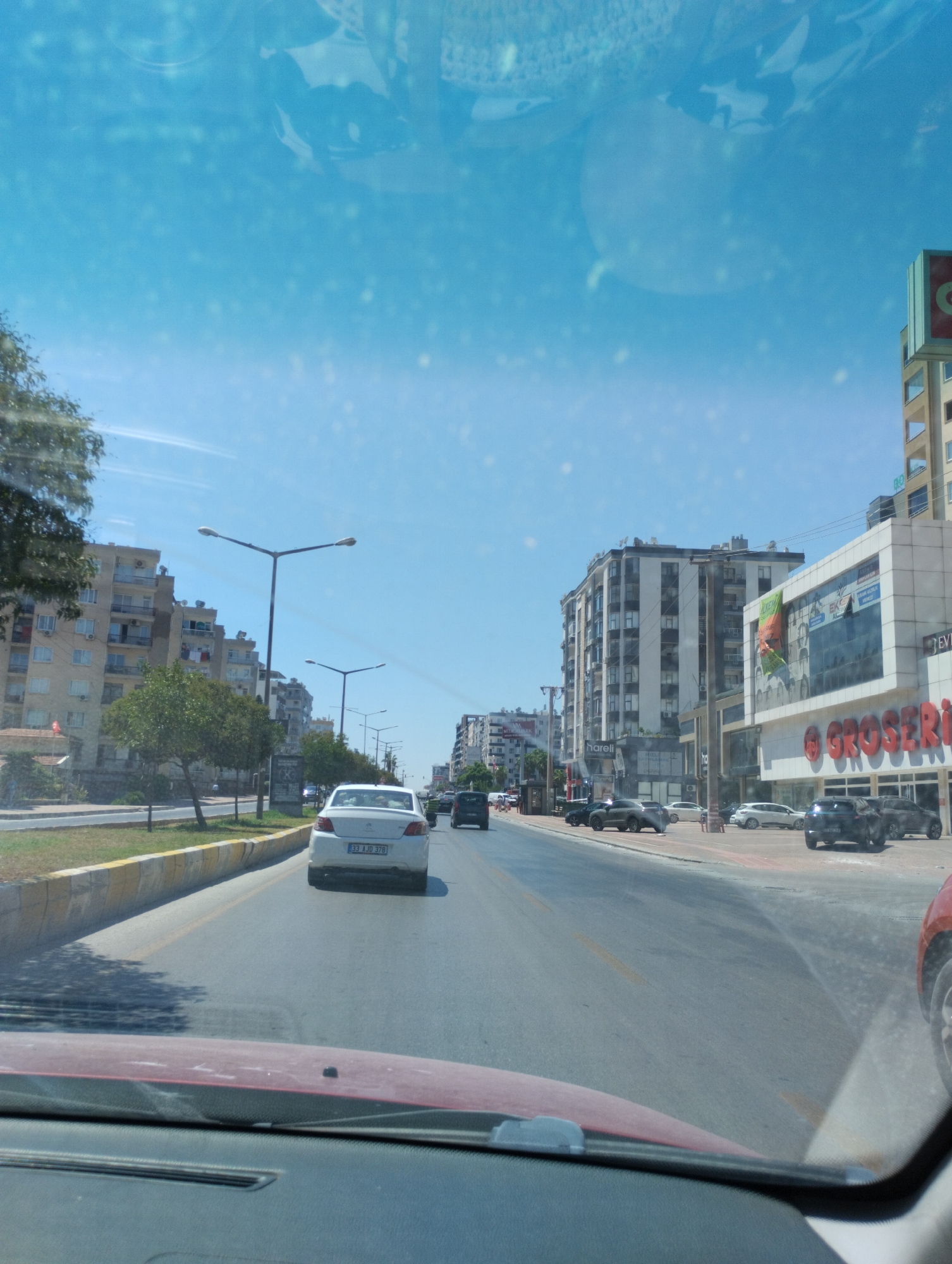 This is a street-level view from inside a vehicle looking forward on a sunny day. The perspective suggests the vehicle is moving in traffic on a multi-lane road.
In the foreground, the red dashboard of the vehicle is visible. The road ahead is paved with asphalt, marked with yellow lane dividers. A yellow and concrete barrier separates the road from a sidewalk and green space on the left.
On the left side of the road, there are several multi-story apartment buildings, some with balconies. Trees are planted along the sidewalk.
On the right side of the road, there are commercial buildings. One building features a large sign that reads "GROSERİ", suggesting a supermarket or grocery store. This building also has a prominent glass facade and advertising posters. Other modern, multi-story buildings line the street.
Several vehicles are visible on the road. A white sedan with license plate "33 AJD 378" is in front of the camera vehicle, traveling in the same direction. Other vehicles, including a dark SUV and a red car, are also present. Pedestrians are sparsely visible on the sidewalk.
The sky is clear blue with some reflections and specks visible on the windshield. The overall environment is urban, with a mix of residential and commercial architecture. The specific details about the location in Mezitli, Turkey, are not explicitly visible within the image itself, but the presence of Turkish license plates and potential Turkish language on signage could indicate this location.