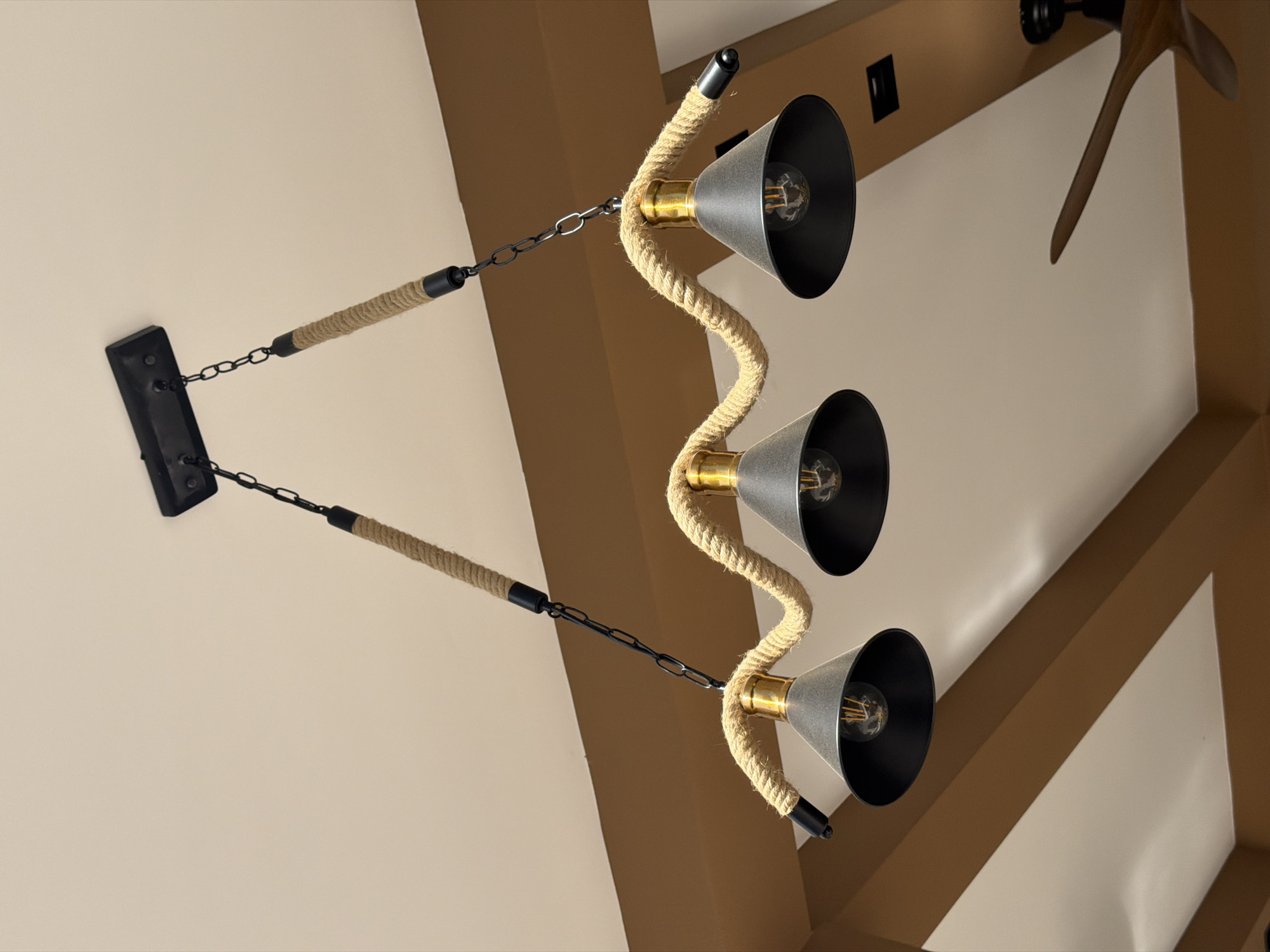 A low-angle indoor shot captures a chandelier with three downward-facing lights. The fixture is primarily composed of a tan-colored rope element. The lights are black with gold bases and are suspended in a wavy pattern along the rope. The lights are illuminated, revealing the light bulbs inside. The rope is connected to the ceiling via black chains and a black mounting plate. Above and slightly to the right of the chandelier, part of a ceiling fan is visible, with blades extending into the frame. The ceiling is painted in a neutral tone. The location is not provided in the image.