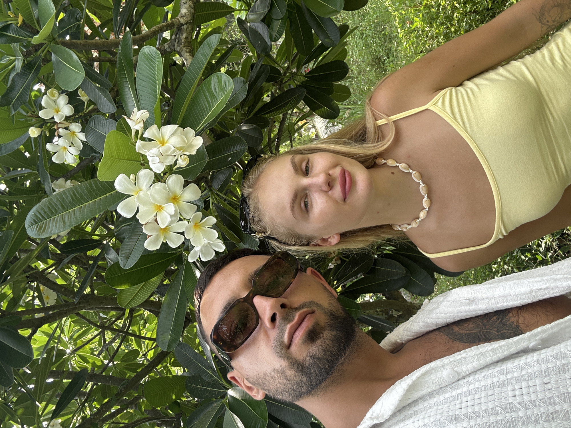 A man and a woman are taking a selfie outdoors, with a flowering tree providing a natural backdrop. The man, on the left, is wearing sunglasses and a white textured shirt. He has a beard and a tattoo visible on his arm. The woman, on the right, has blonde hair and is wearing a yellow tank top and a shell necklace. They are positioned closely together, suggesting they are a couple. The environment appears lush and green, with sunlight filtering through the leaves. The scene is likely set in a tropical or subtropical region, possibly near the city of Nam Nha Trang Ward, Vietnam, given the presence of tropical foliage and flowers.