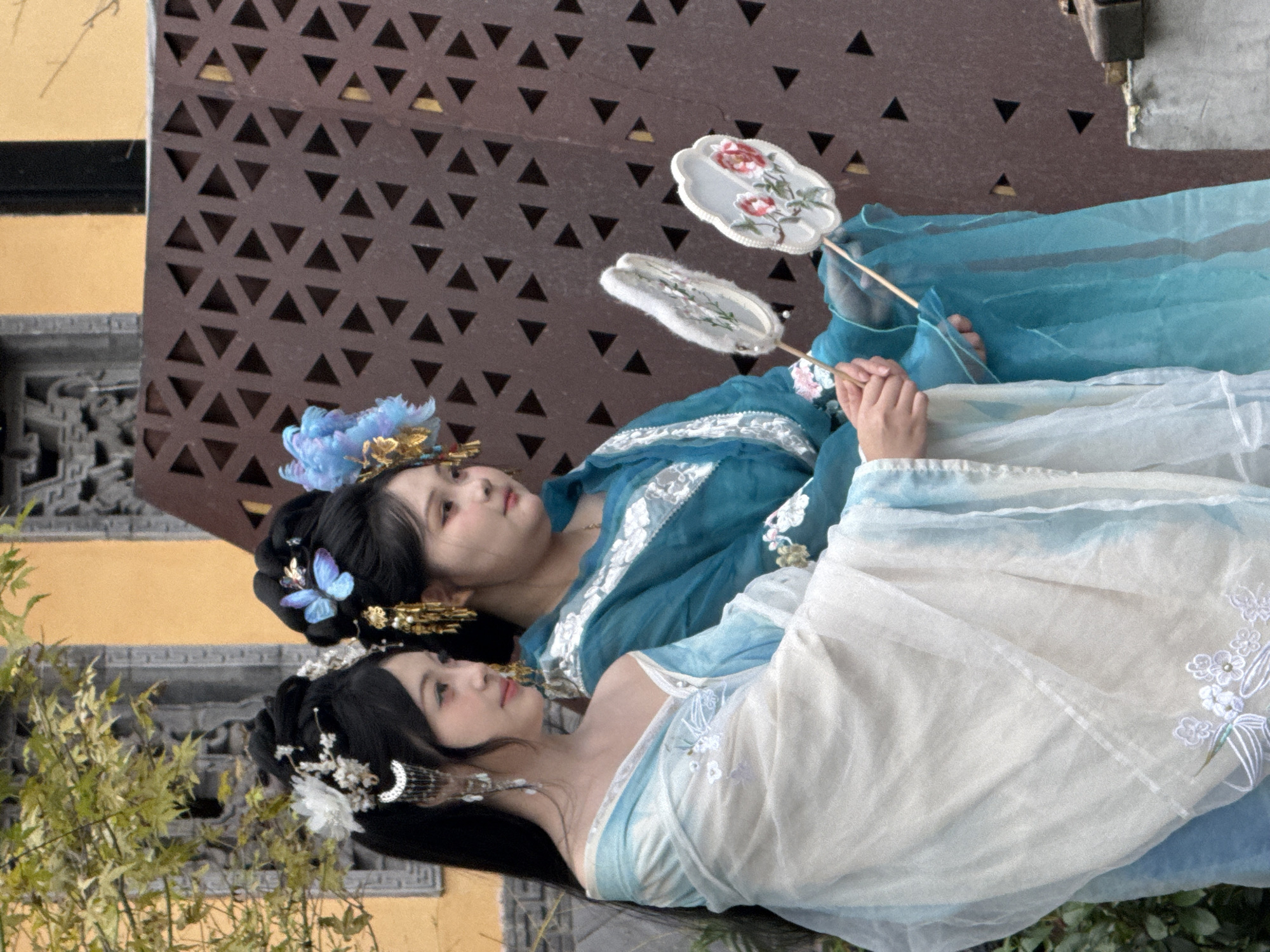 Two young women in traditional Chinese attire are posing for a photo in Yuzhong District, China. The woman on the left wears a light blue and white Hanfu with intricate floral embroidery. Her hair is styled in an updo adorned with white flowers and dangling silver accessories. She holds a small, white fan with embroidered flowers. The woman on the right wears a teal Hanfu with white trim, also with floral embellishments. Her hair is styled similarly, with a blue butterfly hairpiece and gold accents. She also holds a white fan with embroidered flowers. They are standing in front of a decorative, patterned brown screen and a yellow building. Green foliage is visible to the left of the frame.