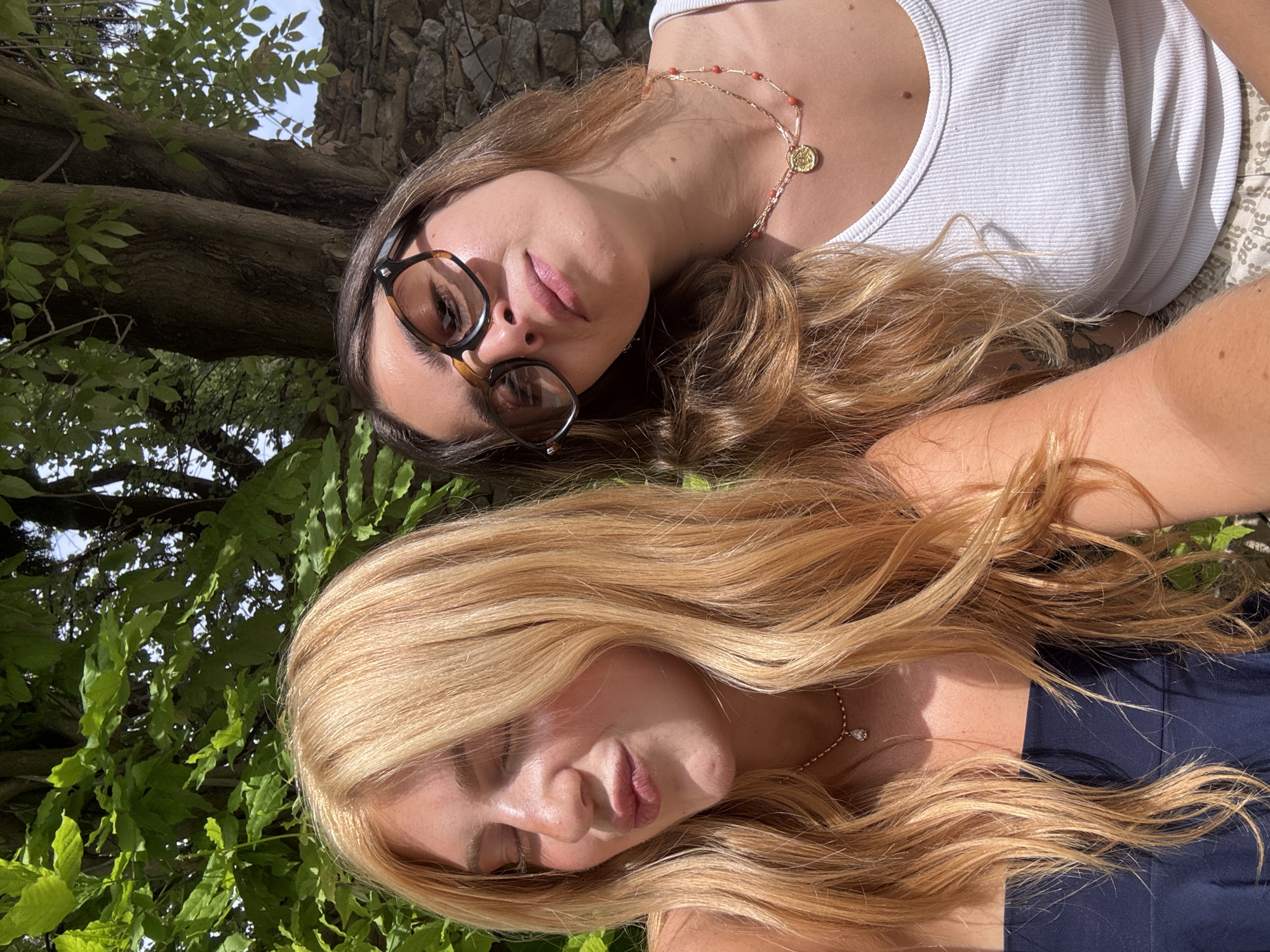 Two women lie supine outdoors in Saint-Victor-la-Coste, France, with a tree trunk and foliage visible above. The woman closer to the top of the image wears glasses, a white tank top, and a beaded necklace. The other woman has blonde hair and wears a navy garment and a delicate necklace. The background includes a stone structure.