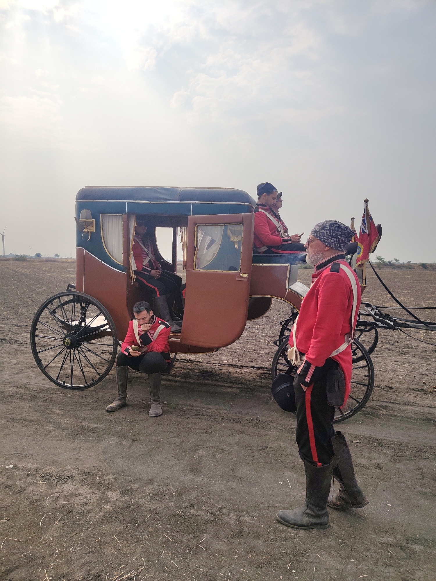 The image depicts an open-air scene in Kottalapalli, India, featuring a horse-drawn carriage and four men dressed in historical-style uniforms. The setting is an unpaved, dry earthen area with sparse vegetation. The background reveals a flat horizon with two wind turbines visible on the far left. The sky is overcast.
A two-toned carriage, primarily brown with dark teal paneling and a blue canvas roof, stands centrally. It possesses large, black spoked wheels and a visible front harness pole. The passenger door on the viewer's left is open. Two flags, a British Union Jack and a plain red banner, are affixed to the rear right side of the carriage.
Four men are present, all uniformly dressed in red tunics with black collars and white cross belts, black trousers with a vertical red stripe, dark knee-high boots, and dark head coverings.
1. **Foreground Right:** A bearded man stands in profile, facing left, wearing a dark patterned beanie. He exhales visible smoke from his mouth and holds a dark, possibly helmet-like, object under his right arm. His left hand is in his pocket.
2. **Left, Seated on Carriage Step:** A man with a beard is seated on the lower entrance step of the carriage, intently focused on and operating a smartphone held in both hands.
3. **Inside Carriage, Left:** A man is seated inside the carriage, visible through the open door, also looking down at what appears to be a smartphone in his hands.
4. **Inside Carriage, Right:** A man is seated on the elevated rear section of the carriage, looking towards the left.
The individuals are not actively interacting with each other; their actions are self-contained. The scene suggests a pause or break in an activity.