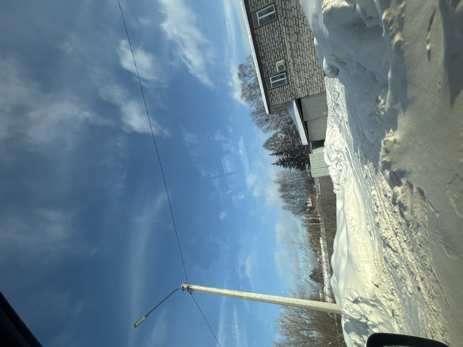 A winter scene with a bright blue sky and wispy clouds. A snow-covered street is on the right side of the image. A building with a brick facade and a couple of windows is set back from the road. A row of trees divides the sky and the snow. A utility pole and a street light are also visible.