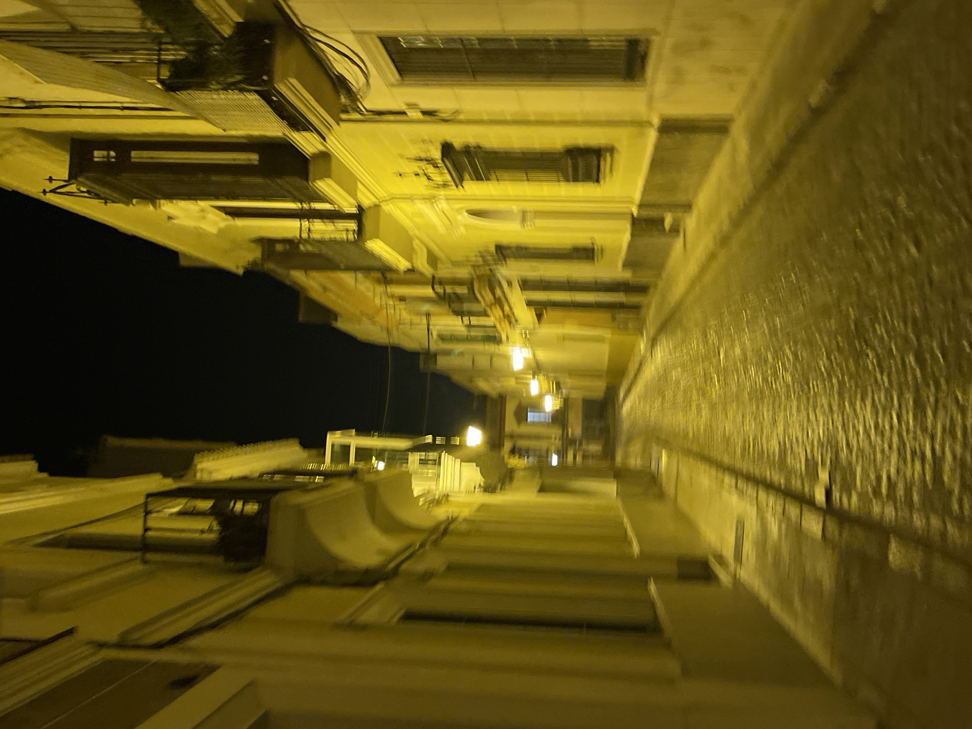 Here is a description of the image:
The image shows a narrow street in Jerez De La Frontera, Spain, at night. The buildings on either side are illuminated with a yellow light, creating long shadows and a sense of depth. The street appears to be paved with stone. The camera is tilted at an angle, giving a distorted perspective. There are no people visible in the image. The buildings are multi-story and have architectural details such as balconies and windows. The sky above is dark.