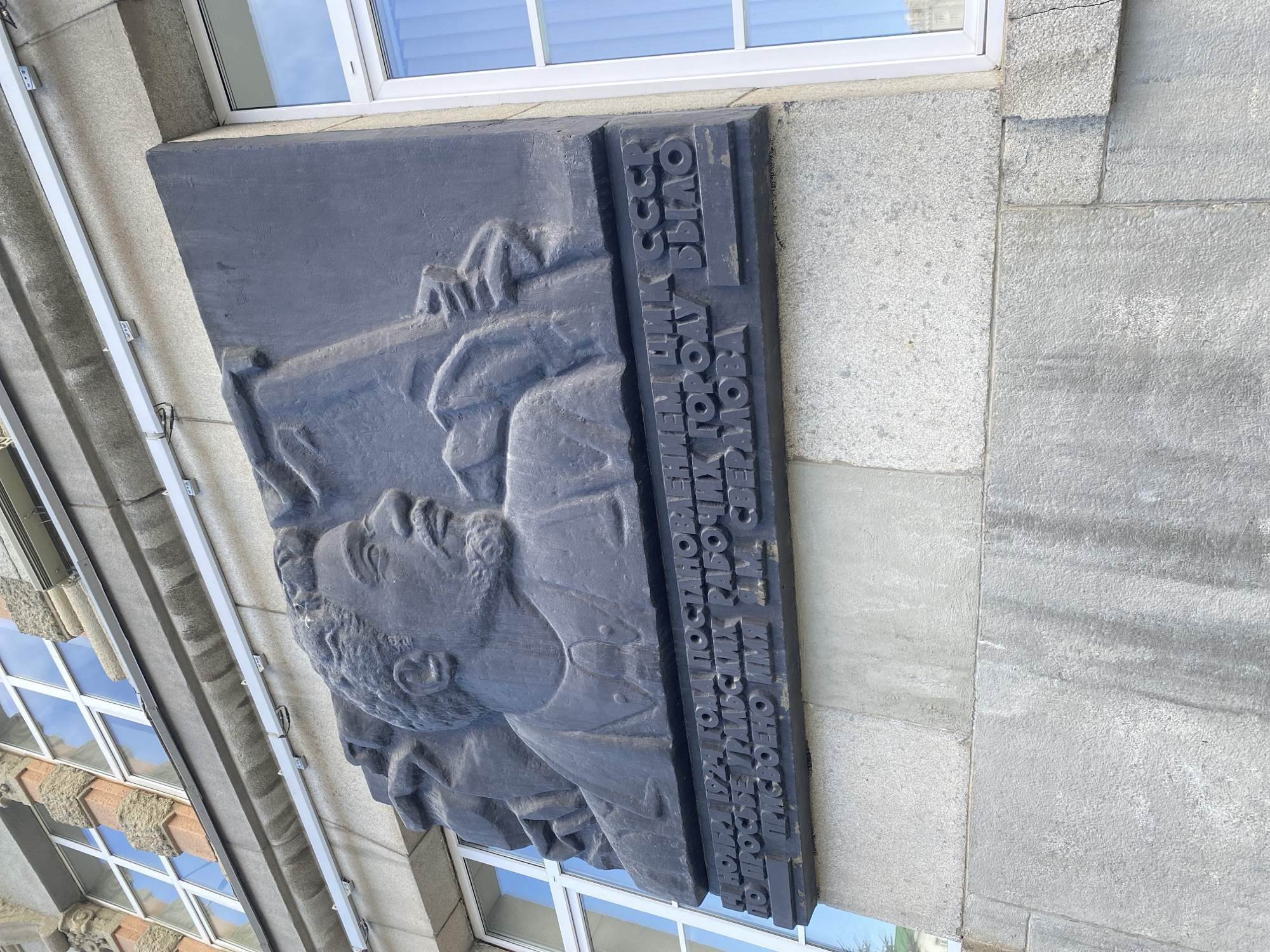 This image depicts a dark-colored memorial plaque affixed to the exterior wall of a building. The plaque features a relief portrait of a man and inscribed Russian text. The text on the plaque translates to: "October 1924 by the decree of the Central Executive Committee of the USSR at the request of the suburban workers, the city was named after Sverdlov." This indicates that the plaque commemorates the renaming of the city, likely Yekaterinburg, to Sverdlovsk, after the Bolshevik leader Yakov Sverdlov. The building exterior appears to be made of gray stone. Reflected in the windows of the building are sky and clouds. The image appears to be taken in Yekaterinburg, Russia.