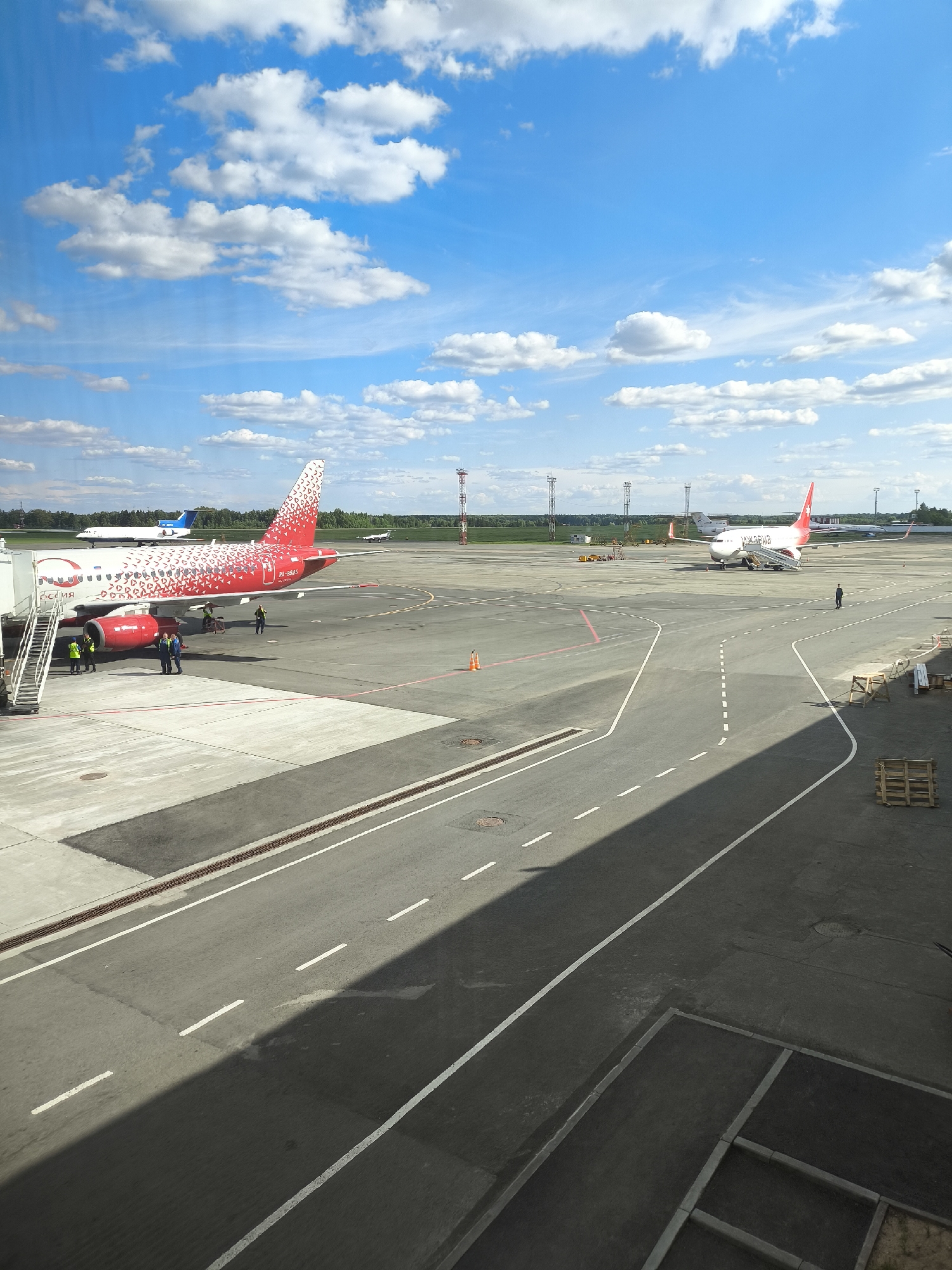The image presents an elevated, outdoor view of an active airport tarmac under a bright blue sky with scattered white cumulus clouds. In the mid-ground on the left, a red and white passenger aircraft is parked, displaying "РОССИЯ" (Russia) on its fuselage and the registration "RA-89045" near the tail. A mobile passenger stair is positioned at its front door, and approximately five to seven ground crew members, wearing high-visibility vests, are attending to the aircraft.
Further into the mid-ground on the right, a second red and white passenger jet, marked with "УРАЛ" (Ural) on its fuselage, is also parked with a mobile stair at its forward door. A single individual, dressed in dark clothing, stands on the tarmac near this aircraft. In the distant background, a blue and white aircraft is visible on the left, and another white aircraft is partially seen on the far right.
The tarmac features white dashed and solid lines, as well as red markings, delineating pathways and operational areas. Several red and white striped light or antenna towers are present in the mid-distance across the field beyond the tarmac. The horizon shows a line of green trees and fields under the clear sky. In the immediate foreground, a dark shadow from the viewpoint structure covers a significant portion of the lower right. Wooden pallets and what appear to be construction materials are stacked in the bottom right corner. The scene depicts typical airport ground operations. The precise location within the city of Unknown, Unknown cannot be determined from the image.