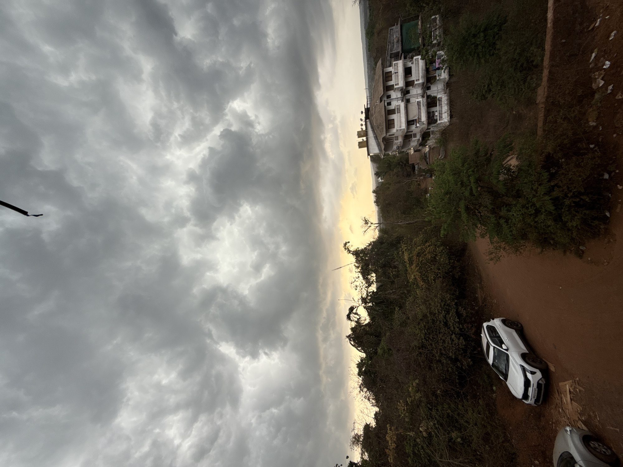 The photograph depicts an overcast sky dominating the view, transitioning into a strip of horizon with a faint glow. Below, the scene transitions into trees and foliage lining what appears to be a brown, dirt-covered path or road. A white SUV, seemingly parked or pulled over, sits on this path, facing toward the upper right corner of the image. To the upper right, a building complex is visible among the vegetation. The photo depicts a location in Sangolda, India.