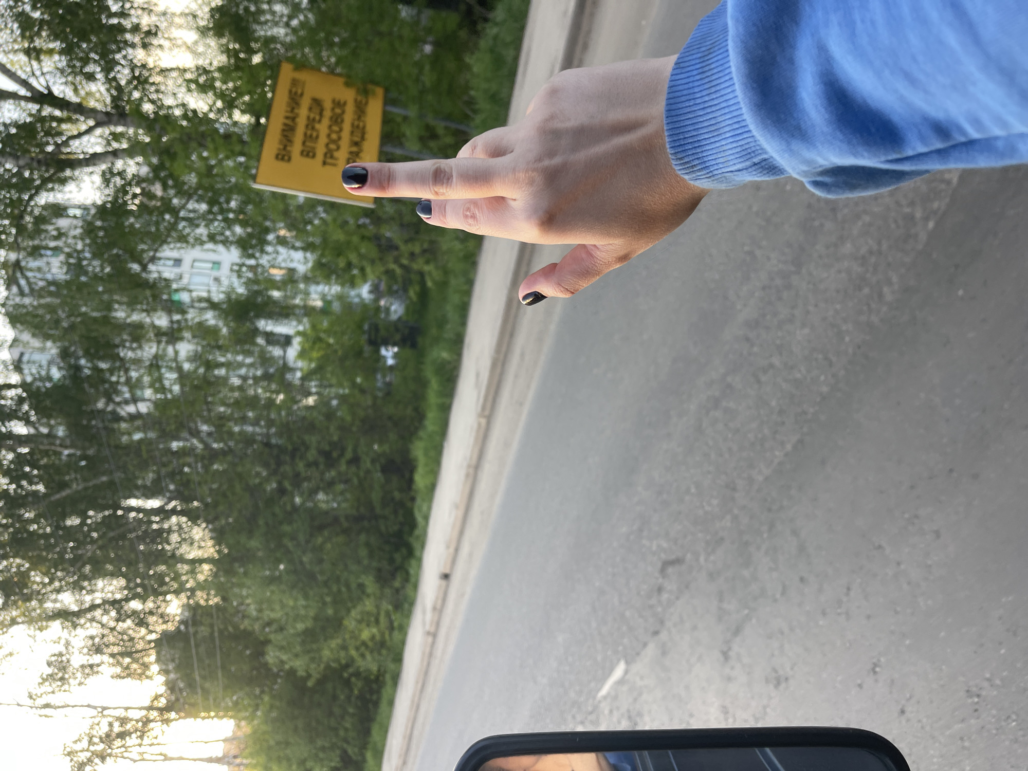 A hand with black nail polish is extended out of a car window, pointing to a yellow road sign. The sign reads "ВНИМАНИЕ! ВПЕРЕДИ ТРОСОВОЕ ОГРАЖДЕНИЕ" in Russian, which translates to "ATTENTION! CABLE BARRIER AHEAD." The scene appears to be in a rural or suburban area, with trees and possibly buildings visible in the background. The road is paved and stretches into the distance. Part of a car's rearview mirror is seen at the bottom of the frame. It is in Molokovo, Russia.