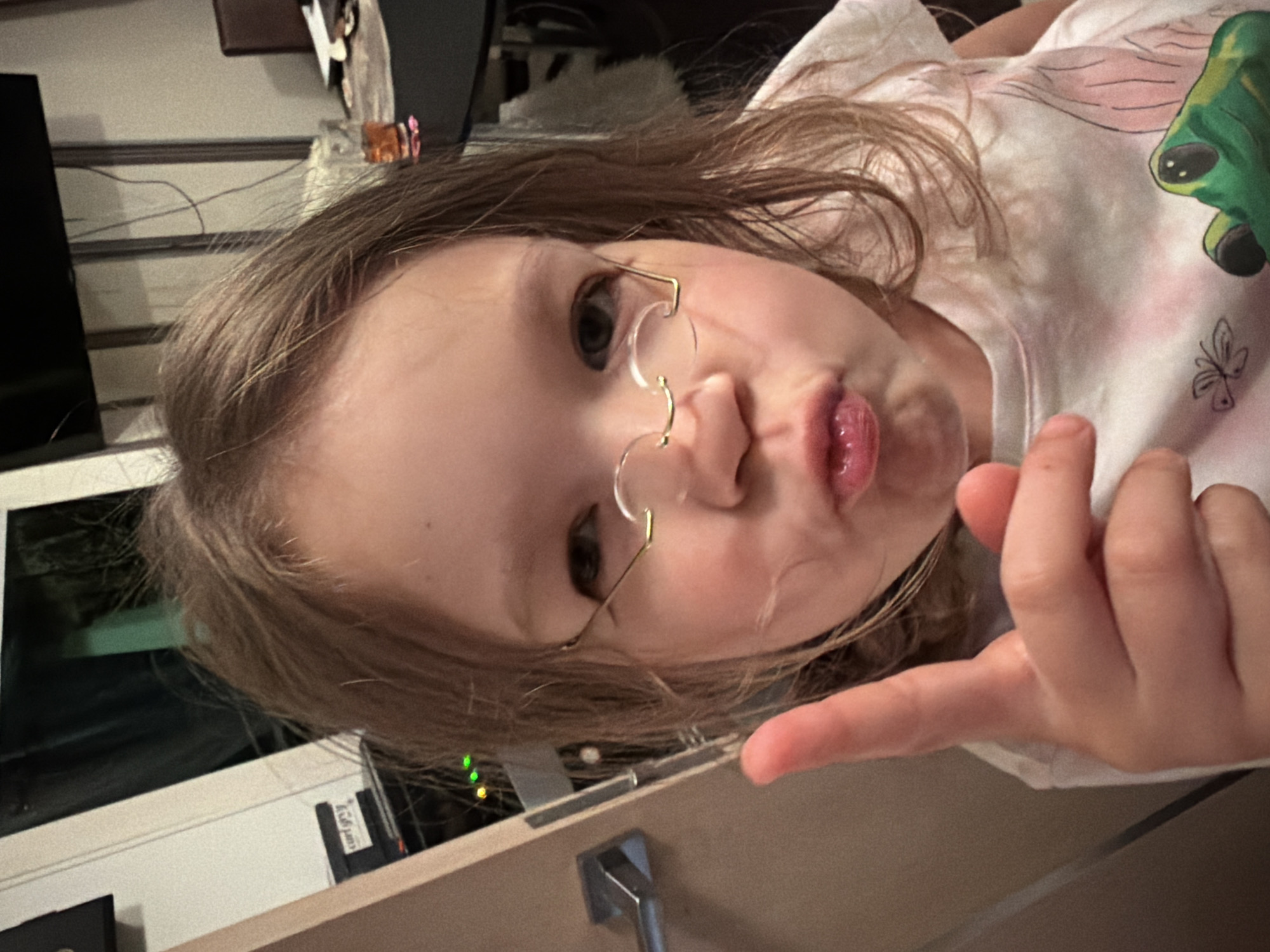 A young girl is positioned close to the camera, wearing rimless glasses and making a pouting face while pointing with her right hand. She is wearing a white t-shirt. The background includes a television, a table, and some household objects. The location is not discernible as being in the city of Saint Petersburg, Russia or any other geographical location from the image alone.