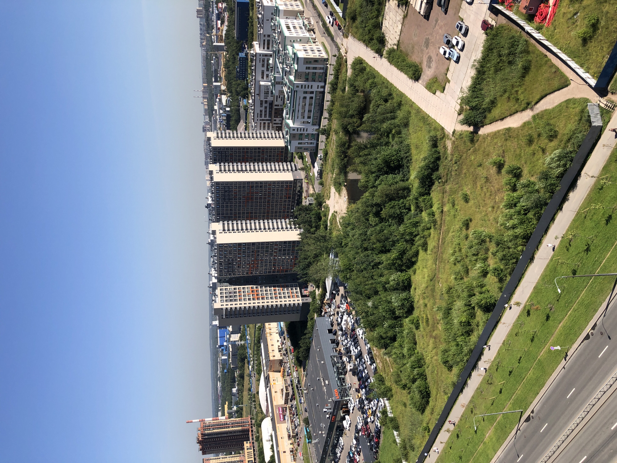 An aerial, high-angle, daylight, outdoor, medium shot shows a sprawling urban landscape under a clear blue sky. In the foreground, a wide asphalt road with white lane markings runs diagonally from the bottom left upwards. To the right of the road is a strip of green grass, followed by a paved pedestrian walkway lined with young trees and streetlights. A black fence runs along the far edge of the walkway, separating it from a large, uneven field of green grass and scattered trees.
Further into the scene, a group of modern, multi-story apartment buildings dominate the mid-ground. Several of these buildings are tall and slender, with a repeating pattern of windows and balconies. Some have a light-colored facade with darker accents. To the left of these buildings, a long, low-rise commercial building with a light-colored facade is visible, with a parking lot full of cars in front of it. A car dealership with a prominent "LADA" sign is also present, with many cars parked in its lot.
Beyond the apartment buildings and commercial areas, the urban landscape extends into the distance, with more buildings, industrial structures, and green spaces visible under the hazy sky. Two tall, cylindrical smokestacks rise in the far distance. The overall scene depicts a mixed-use urban environment with residential, commercial, and recreational areas.