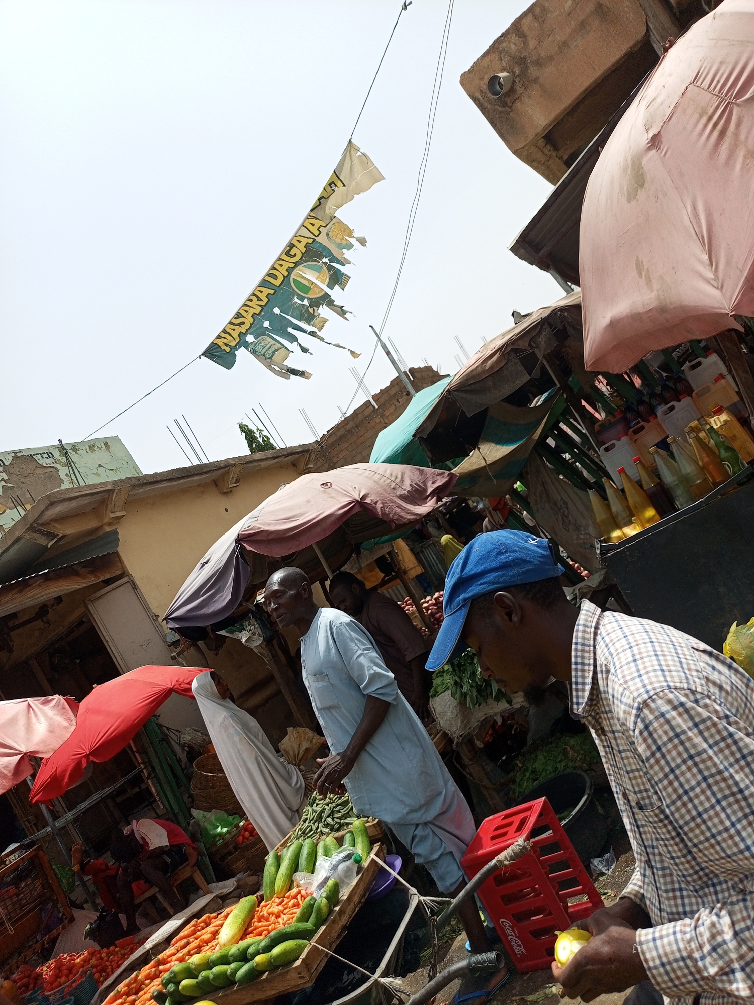 The image depicts a bustling marketplace in Arawa, Nigeria. Several individuals are present, including men in traditional attire and casual clothing. One man in a blue cap and plaid shirt is peeling a lemon. Produce is displayed in wooden crates, including tomatoes, carrots, and cucumbers. Overhanging the market stalls are colorful umbrellas and a tattered banner with the words "NASARA DAGAA" visible. The environment is crowded with various goods, including bottles of oil and other merchandise, creating a lively and active scene.