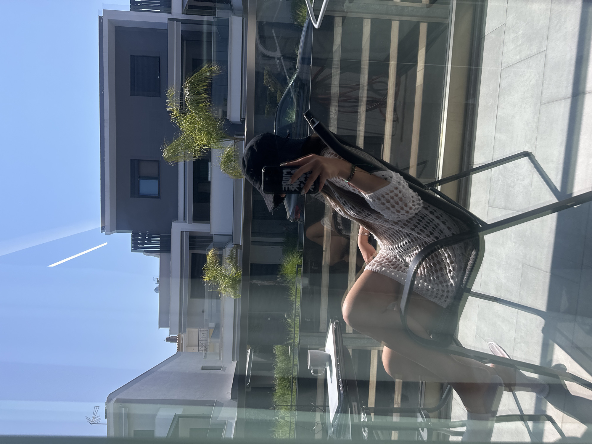 A woman is seated in a black folding chair on a balcony, holding a smartphone in her right hand and a white mug on a small table to her left. She is wearing a white, crocheted cover-up, a black bucket hat, and pink sandals. The balcony overlooks a street with modern buildings, green vegetation, and a parked black car. The scene is illuminated by bright sunlight, casting shadows on the tiled floor. The woman's face is obscured by the phone.