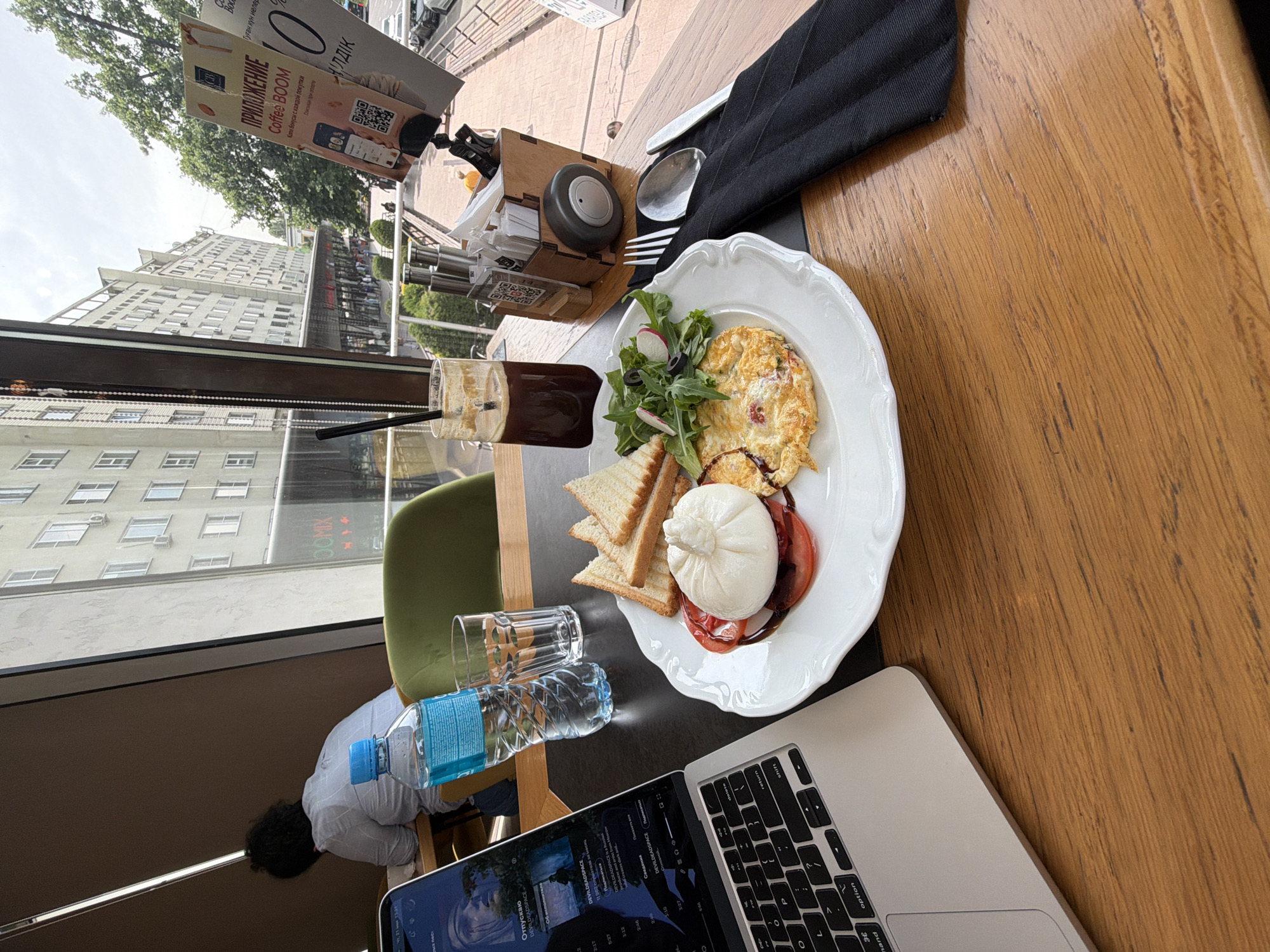 A first-person view shows a table setting indoors, likely at a café or restaurant in Almaty, Kazakhstan. A partial view of a silver laptop sits at the bottom right corner of the frame, partially obscuring a bottle of water and a clear glass. The table is partially occupied by an oval white plate containing an omelet, salad with radishes, slices of tomato with burrata, and toasted bread. A tall glass of dark liquid, likely coffee, sits next to the plate. Cutlery wrapped in a black napkin rests on a wooden surface at the top right, alongside a wooden container with cards, condiments, and a gray weight.
A man is seated in the background to the left, wearing a light-colored shirt. Beyond the man, the café window shows a view of a building across the street with an advertisement for “Coffee BOOM” displayed on the glass. The street and surrounding buildings are visible through the window.