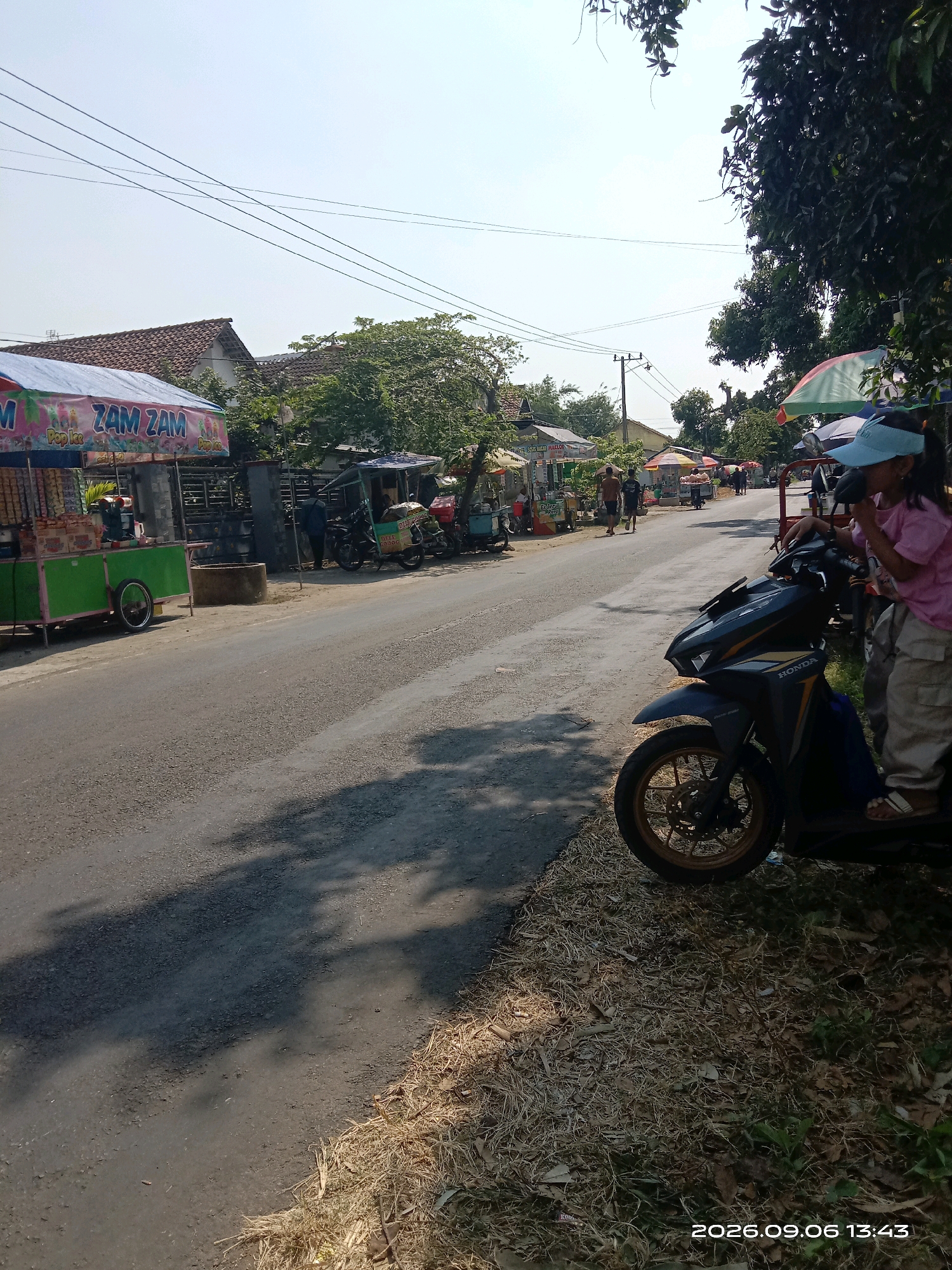 The image captures a street scene in Sepanyul, Indonesia, on a sunny day. In the foreground, a dark-colored Honda scooter is parked on the side of a paved road. A young girl wearing a pink shirt and light-colored pants, with a light blue cap on her head, is seated on the scooter, appearing to be eating something. Her feet are visible, wearing light-colored sandals. To the left of the scooter, dry grass and leaves are scattered along the roadside.
The road stretches into the background, lined with various roadside stalls and small shops. On the left side of the road, a bright green food cart with a blue and pink awning is prominently displayed, featuring the text "ZAM ZAM" and "Pop Ice". The cart contains packaged goods. Further down the road, other stalls with colorful umbrellas are visible, with people and motorcycles in the vicinity. In the background, houses with tiled roofs and trees can be seen under a clear blue sky with scattered power lines. A date and time stamp at the bottom right corner reads "2026.09.06 13:43". The overall environment suggests a local marketplace or a busy street with informal vendors.