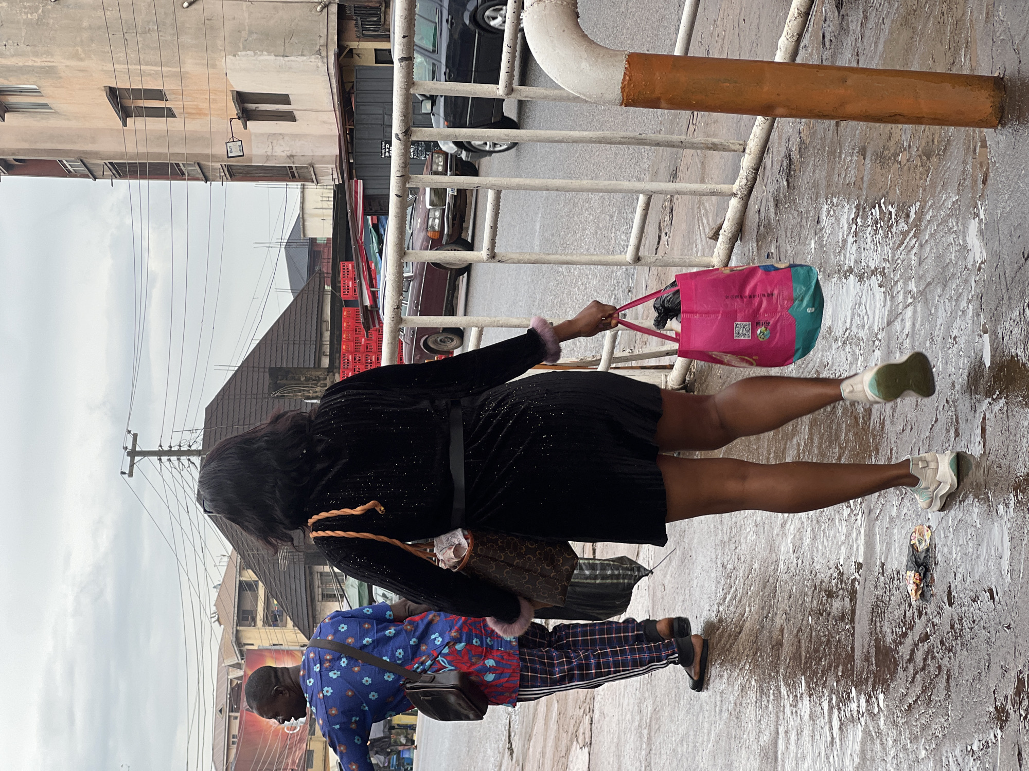 This media file depicts two people walking on a wet, paved street in Nkpor, Nigeria. The person in the foreground is a woman wearing a black, sparkly dress, white sneakers, and carrying a pink tote bag over her shoulder. She has long, dark hair and appears to be walking away from the camera. The second person is a man walking in front of her, wearing a blue shirt with a floral pattern and plaid pants. He is carrying a dark bag over his shoulder and is also walking away from the camera.
The scene appears to be a street in a commercial area, with buildings and a fence visible in the background. Red plastic crates are stacked on the side of the street, suggesting a marketplace or distribution point. Several cars are also visible on the road, including a dark-colored sedan and a burgundy Mercedes-Benz. The ground is wet, indicating recent rain, and the sky is overcast, suggesting a cloudy or potentially rainy day. The overall atmosphere seems to be that of daily life on a busy street. There are no explicit signs of time of day or specific emotions, beyond the general impression of movement and activity. No readable text is visible in the media file.