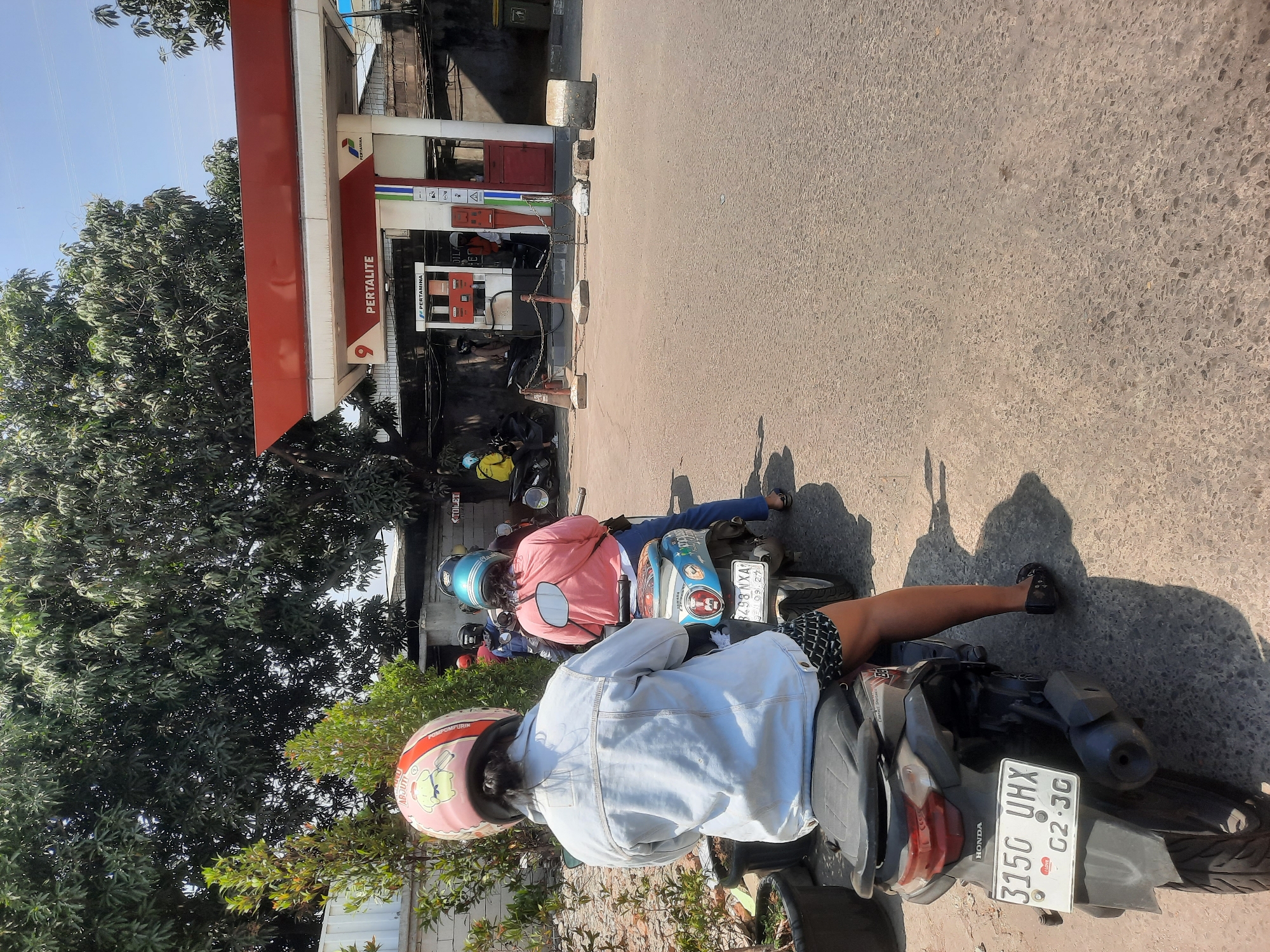 In North Jakarta, Indonesia, a sunny daytime scene unfolds at a Pertamina gas station. The foreground features the rear of a dark-colored scooter with a license plate reading "315G UHX G2.30". A person wearing a light blue denim jacket, black patterned shorts, and black sandals sits on the scooter, their back to the camera, with a pink helmet adorned with a cartoon character. Behind them, another scooter is occupied by a person in a pink long-sleeved shirt and dark pants, also wearing a helmet. Further back, more people on scooters are visible near the gas pumps. The gas station itself has a red awning with "PERTAMINA" signage. Trees with dense green foliage line the background, and the sky is clear and blue. The ground is paved with asphalt.