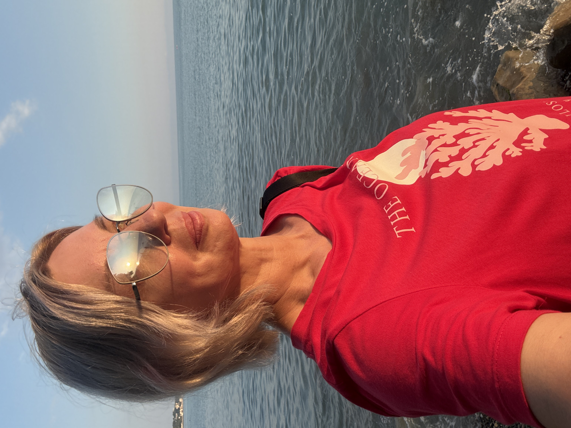 A woman stands by the sea in Sochi, Russia, wearing a red t-shirt with a coral graphic and the words "THE OCEAN" printed on it. Her blonde hair is short, and she wears large, reflective sunglasses. The sun is setting, casting a warm glow on her face and reflecting in her sunglasses. The sea is calm, with gentle waves lapping against the shore, which is made up of stones and rocks. The sky is a clear blue with a few wispy clouds. In the distance, a faint line of buildings can be seen along the coastline. The overall scene depicts a serene moment by the Black Sea at sunset.