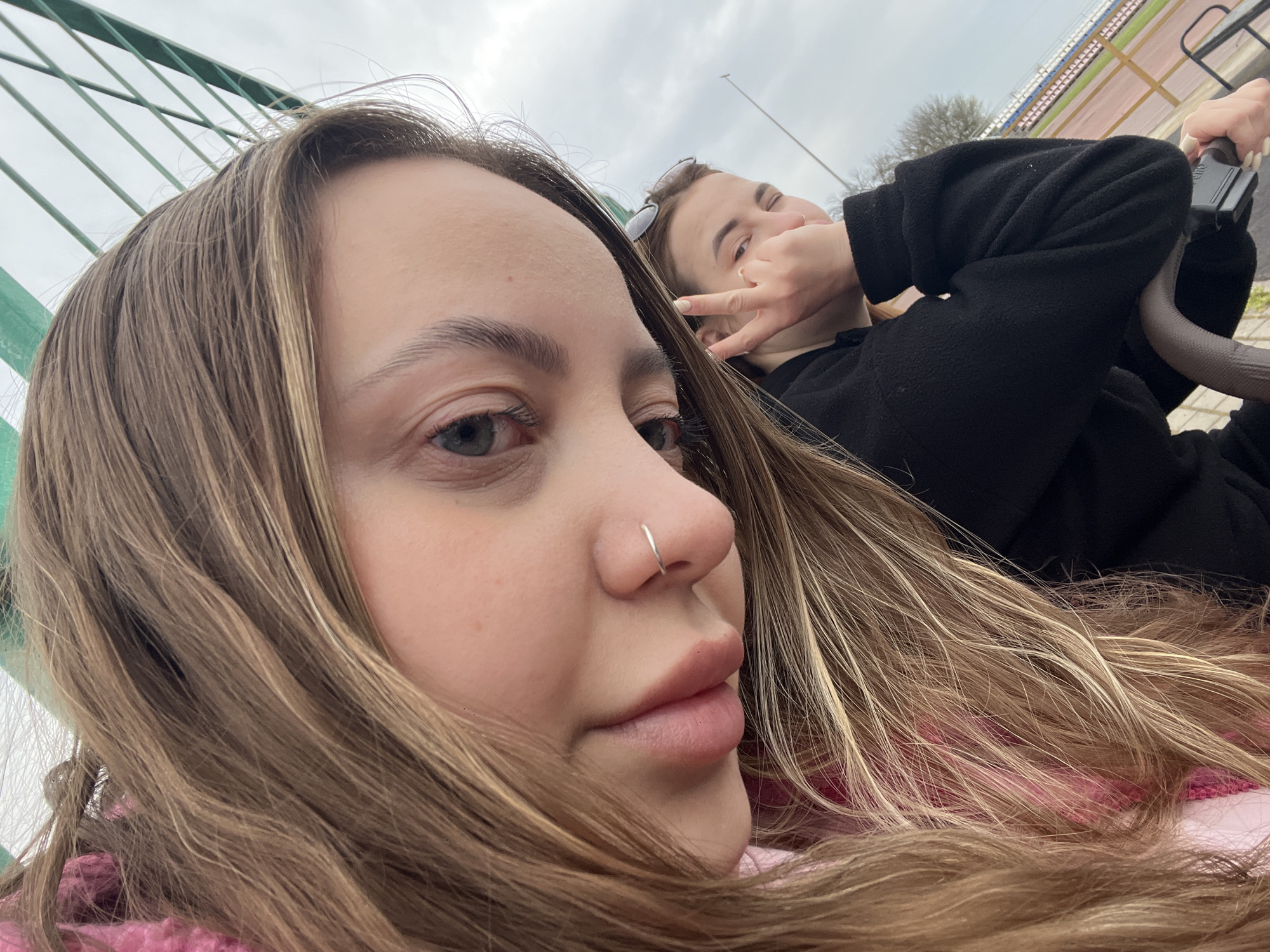 The image features two young women in an outdoor setting, likely a stadium or sports complex in Novopokrovskaya, Russia, on an overcast day.
In the foreground, a woman's face is prominent, looking towards her left. She has long, light brown hair with blonde highlights, a subtle expression, and a small silver nose ring. Her hair partially covers a pink garment worn underneath.
Behind her, a second woman is partially visible, lying down and facing the camera with a playful expression. She wears a black long-sleeved top and has sunglasses pushed up onto her head. Her left hand is raised, forming a "peace" or "V" sign, while her right hand holds onto a dark, curved handle, possibly part of a seating structure or a ride.
The background shows a cloudy sky and, to the right, rows of stadium seating or bleachers. A green metal railing is visible in the upper left, suggesting an enclosure or barrier. The overall atmosphere is casual and relaxed. No visible text is present in the image.