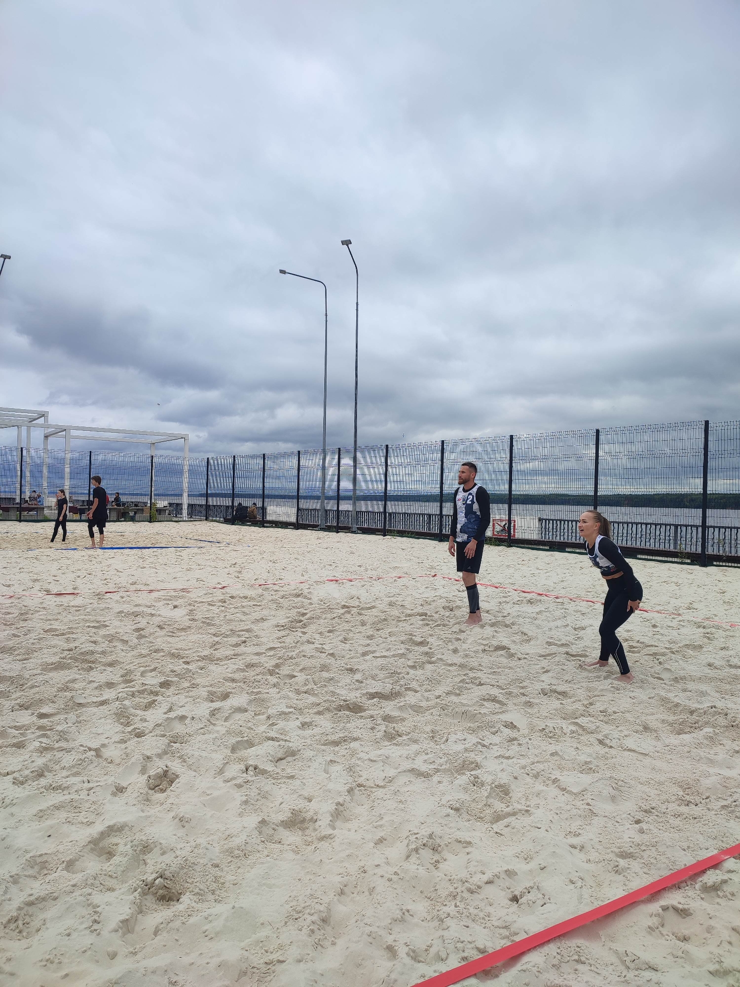 This is a full-shot, eye-level, outdoor photograph taken on a cloudy day at a sandy beach volleyball court. In the foreground, a sandy court with red boundary lines is visible. In the mid-ground, two pairs of people are playing volleyball. On the left, a man and a woman in black athletic wear are visible, facing away from the camera. On the right, a man in a black and white jersey and black shorts, and a woman in black athletic wear are actively engaged in the game. They are positioned on opposite sides of the court. In the background, a black chain-link fence separates the court from a body of water, likely a river or lake. Beyond the water, a hazy shoreline is visible under a heavily overcast sky with visible streetlights. A white canopy structure is also visible to the far left of the frame.