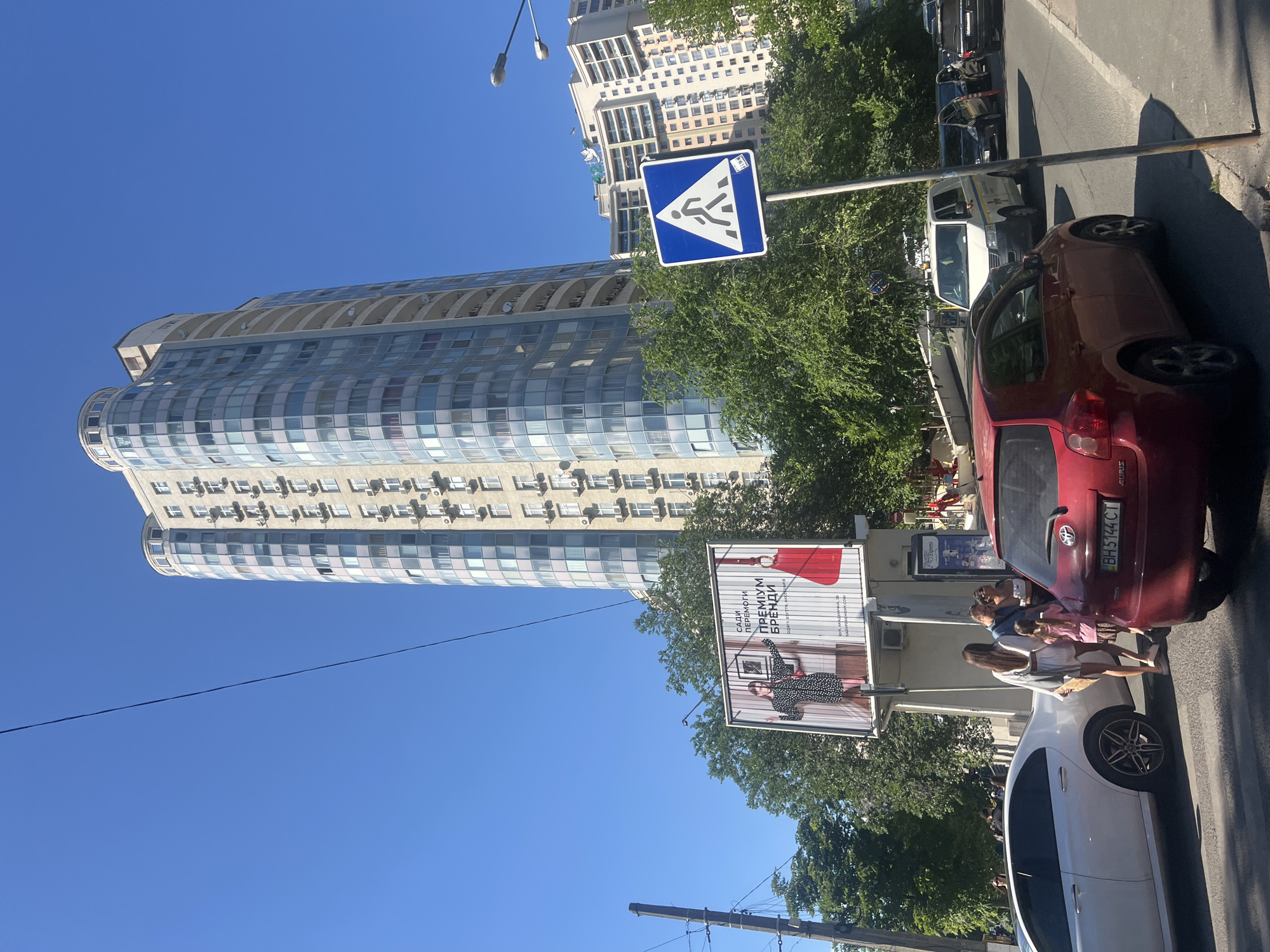 The image depicts a street scene in Odesa, Ukraine, on a clear, sunny day. Dominating the skyline is a tall, modern, curved apartment building with a light-colored facade and numerous balconies and windows. In the foreground, a red hatchback car is stopped at a pedestrian crossing, indicated by a blue and white pedestrian crossing sign with a white pedestrian silhouette on a blue background. To the left of the red car, a white sedan is also stopped.
Several people are visible near the pedestrian crossing. A woman in a white outfit and a young girl in a pink dress are walking away from the red car. Behind them, a group of people, including two women and a child, appear to be crossing the street. A billboard advertising "PREMIUM BRANDS" is situated behind the pedestrian crossing area, featuring a model and a red handbag.
The street is lined with lush green trees, and other buildings, including a lighter-colored apartment complex with arched balconies, are visible in the background. Traffic is present, with several cars lined up on the road, including a white van that appears to be a service vehicle with yellow and blue markings. Power lines and street lamps are also visible in the scene. The overall impression is of an urban environment on a busy day.