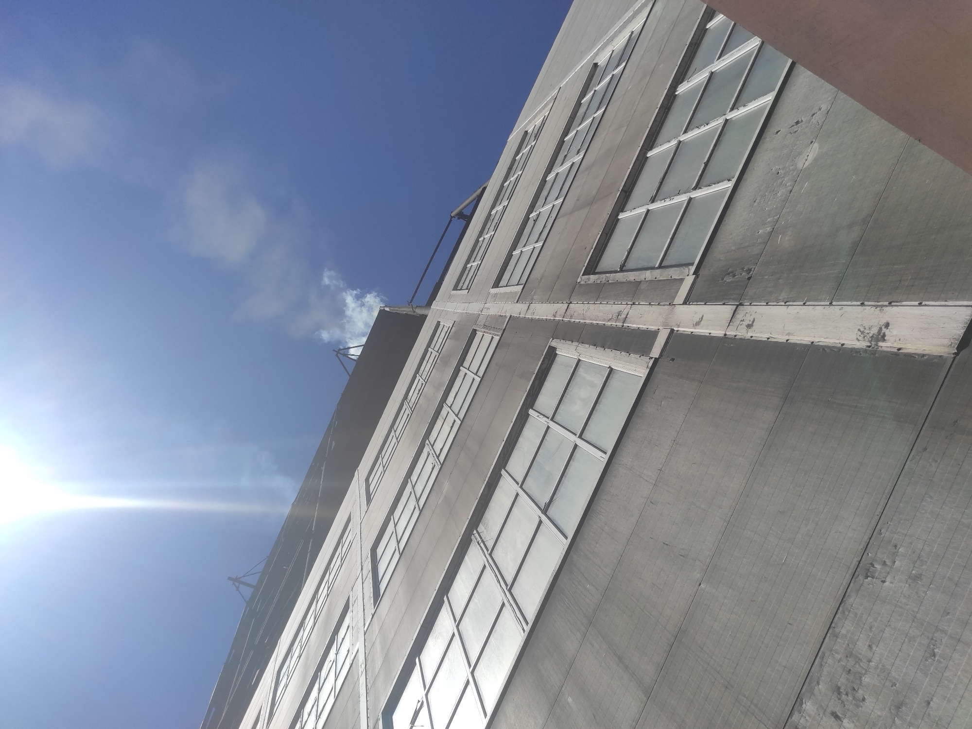 The image is an upward-angle shot of a tall, industrial-looking building with a grey, textured facade and numerous large, multi-paned windows. The building is seen from a low perspective, emphasizing its height against a bright blue sky with some wispy white clouds. The sun is very bright and positioned in the upper left corner, creating lens flare and washing out parts of the sky. A thin plume of white smoke or steam is visible rising from the top of the building against the sky. The building appears to be made of concrete panels, with vertical seams and some signs of wear and tear on the facade. The windows are framed in a light color, possibly white or silver, and reflect the bright sky. The scene appears to be in an urban or industrial setting. There are no people visible in the image. The building's architectural style suggests it could be a factory, warehouse, or older commercial building. The location of Kyzyl, Kazakhstan is not explicitly shown or implied by any visual elements in the image itself, but is provided as context.