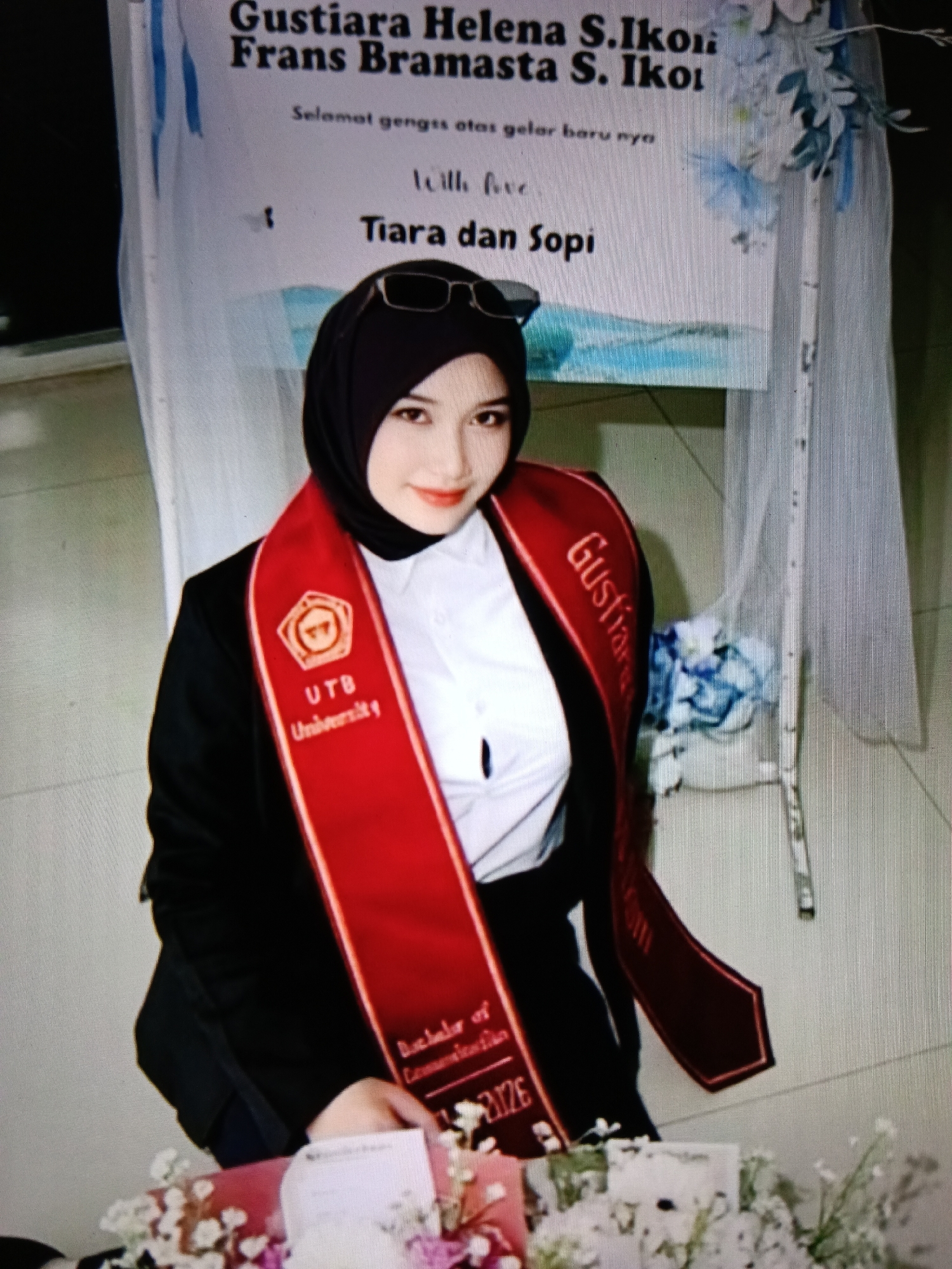 A young woman wearing a black hijab and a black suit with a white shirt and a red sash is posing for a photograph. The sash has a logo with "UTB University" written on it and "Gustiana" is also visible. She is wearing glasses on top of her head. She is standing in front of a banner with text, some of which is visible: "Gustiana Helena S. Ikoni Frans Bramasta S. Ikoi", "Selamat gengs atas gelar baru nya", and "Tiara dan Sopi". There are flowers in the foreground and on the right side of the image. The background appears to be an indoor setting, possibly a graduation ceremony. The overall scene suggests a formal event or celebration. This image does not contain any information that definitively places it in Raha, Indonesia.