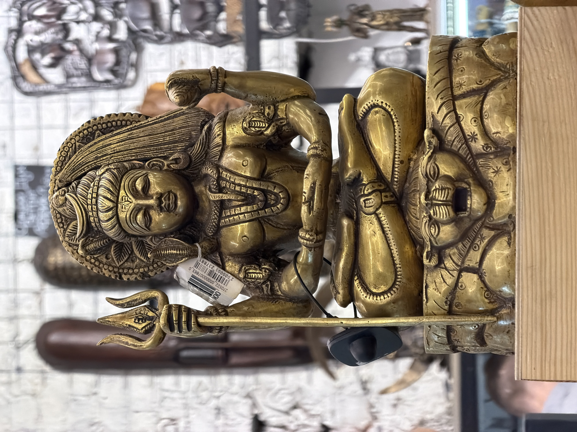The image shows a golden statue of a Hindu deity, likely Shiva, displayed on a shelf or surface, possibly for sale. The statue is intricately detailed, featuring multiple arms, a trident, and a serpent around its body. A security tag and a price tag are attached to the statue, indicating it's in a retail environment.
The background is blurry but appears to show other statues or decorative items, along with a white brick-like wall.
There's no clear indication of Moscow or Russia in the image based solely on the visual content. The statue itself is of Hindu origin, which wouldn't inherently tie it to a specific location like Moscow without additional context.