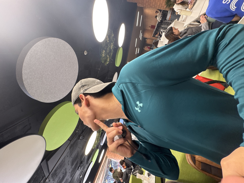 A male subject is prominently featured in the foreground of a modern communal space in Mexico City, Mexico. He wears a grey baseball cap backward and a teal long-sleeved Under Armour quarter-zip shirt. His head is tilted upwards and to the left, with his right index finger placed against his chin, while his right hand holds a small, white, cylindrical device. The ceiling above is dark, adorned with multiple circular acoustic panels in white, light green, and gray, interspersed with circular recessed lighting fixtures. In the mid-ground, green upholstered chairs and white tables are visible, occupied by several other individuals, some engaged with mobile phones. A red brick wall forms part of the background on the right side of the frame. The overall environment suggests a contemporary cafeteria, food court, or educational common area.