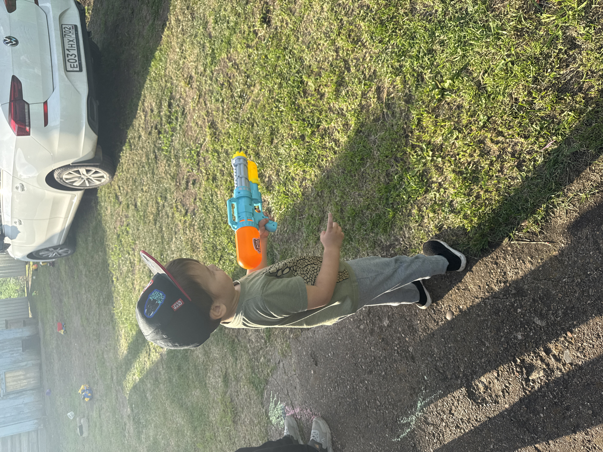 In a rural outdoor setting, likely a backyard in Чуй-Атасево, Russia, a young child, wearing a black baseball cap with Star Wars motifs, a green t-shirt with a dinosaur print, and grey sweatpants, stands on a gravel path, holding a blue and orange toy water gun. The child appears to be in the process of playing, with a stream of water visibly spraying from the gun towards the right side of the frame. In the background, a white Volkswagen sedan is parked on a grassy area. The environment is characterized by a weathered wooden structure to the left, a patch of dirt path, and a sun-drenched lawn with scattered toys, suggesting a residential area. The lighting indicates it is daytime, with long shadows cast by the sun.
