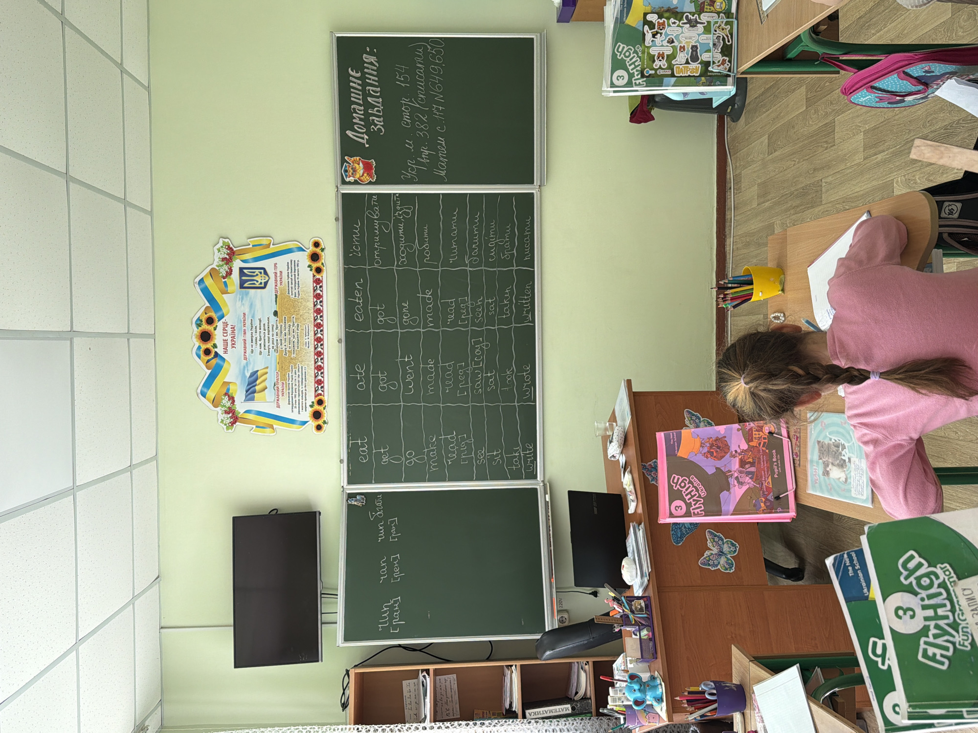 The image shows a classroom in an unidentified location. A young girl sits at a wooden desk, seemingly working on schoolwork. A chalkboard on the wall displays verb conjugations in English and Ukrainian, along with a homework assignment. A poster featuring Ukrainian national symbols hangs above a television mounted on the wall. Books and school supplies are scattered on the desk and shelves nearby. The room appears to be set up for a language lesson.
